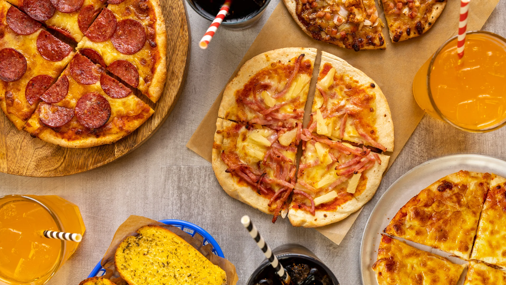 Several pizzas with various toppings, glasses of orange soda with striped straws, and a basket of garlic bread are arranged on a table seen from above.