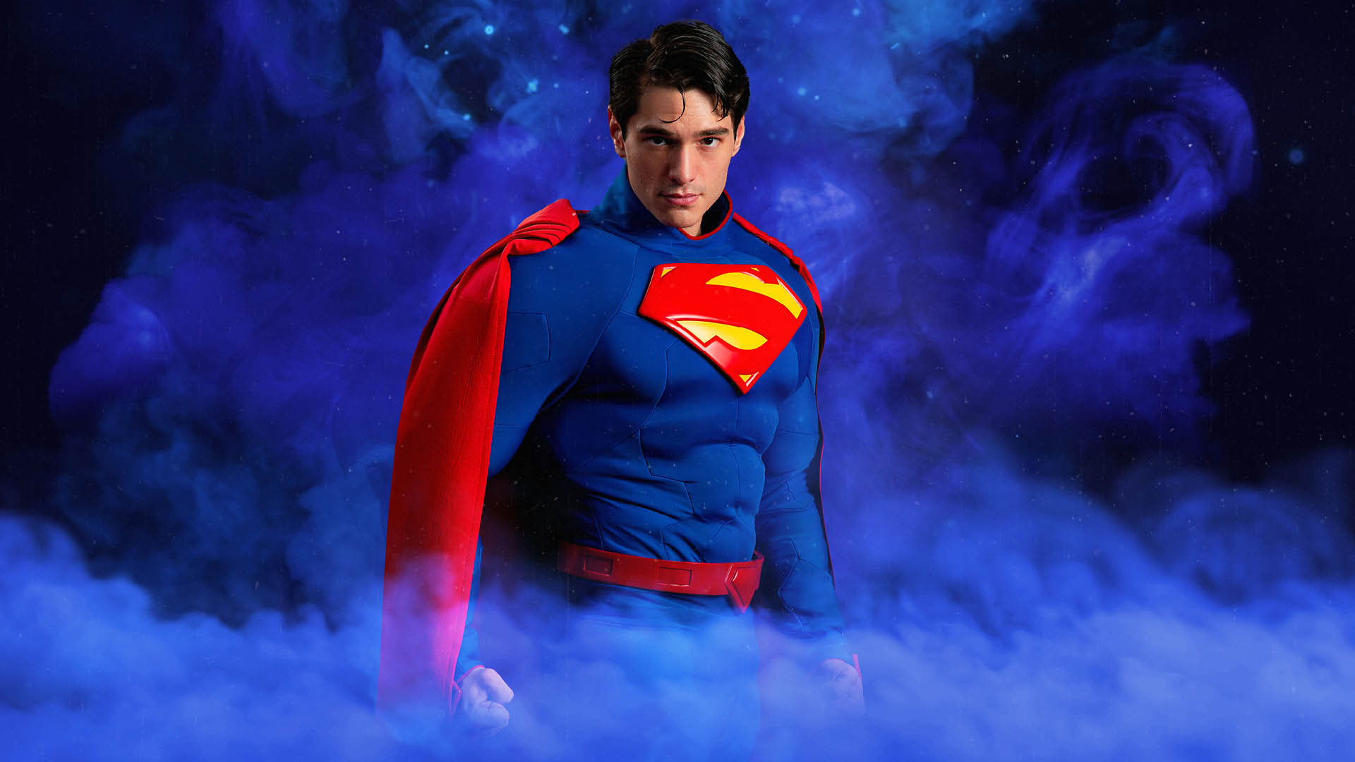 A person dressed as Superman stands in front of a purple, smoky background, wearing a blue suit with a red cape and the Superman logo on the chest.
