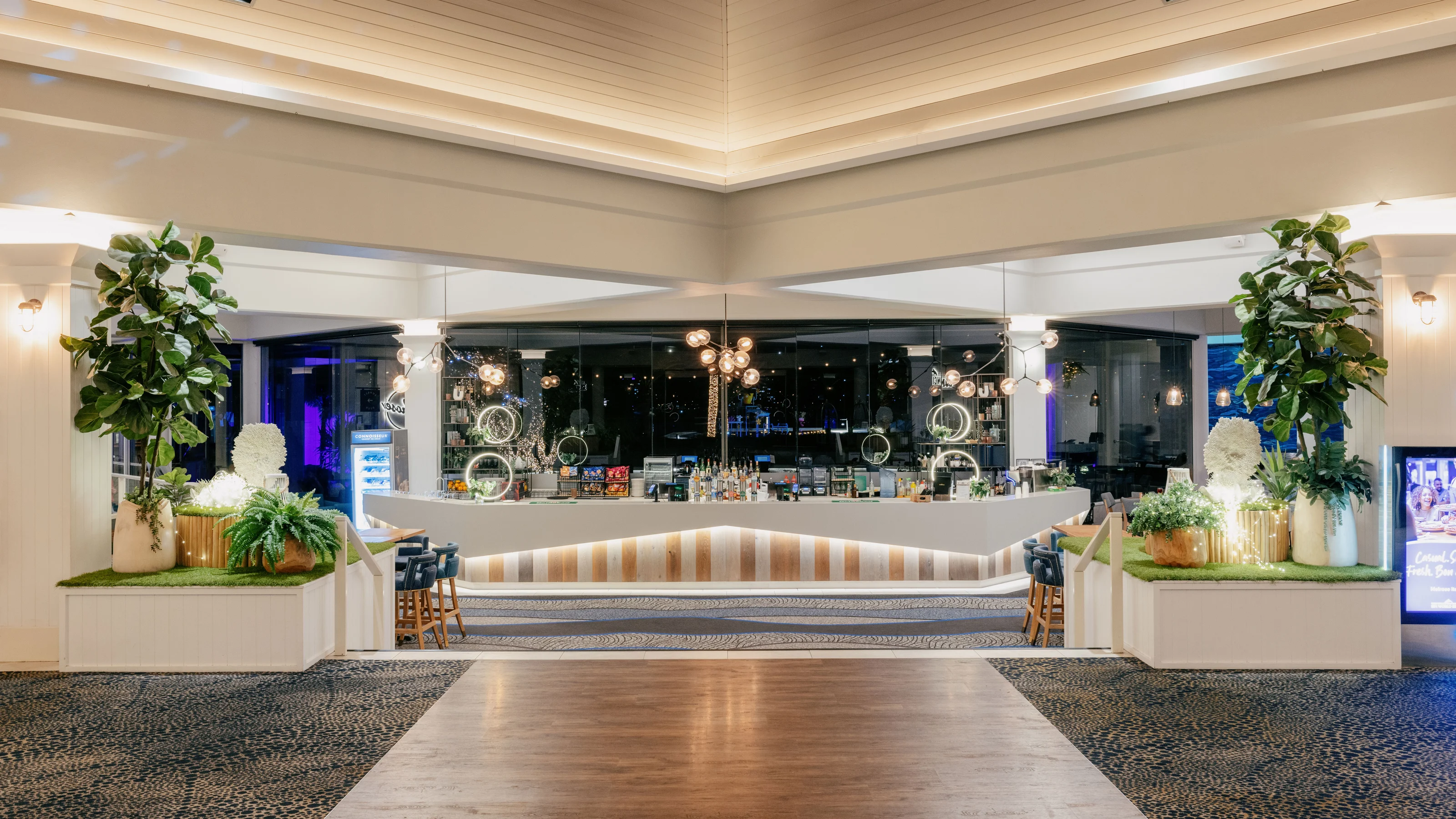 Modern indoor bar (Lobby Lounge at Sea World Resort) with a central counter, decorative lighting, and potted plants. Pathway leading to the bar with tables and chairs on either side. CE