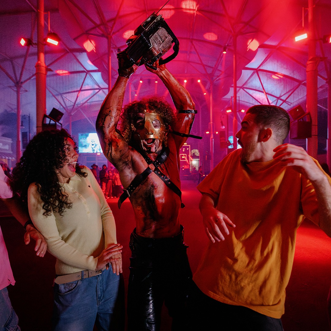 Four startled people react as a performer in a mask and costume wields a chainsaw under dramatic red lighting at an indoor event.