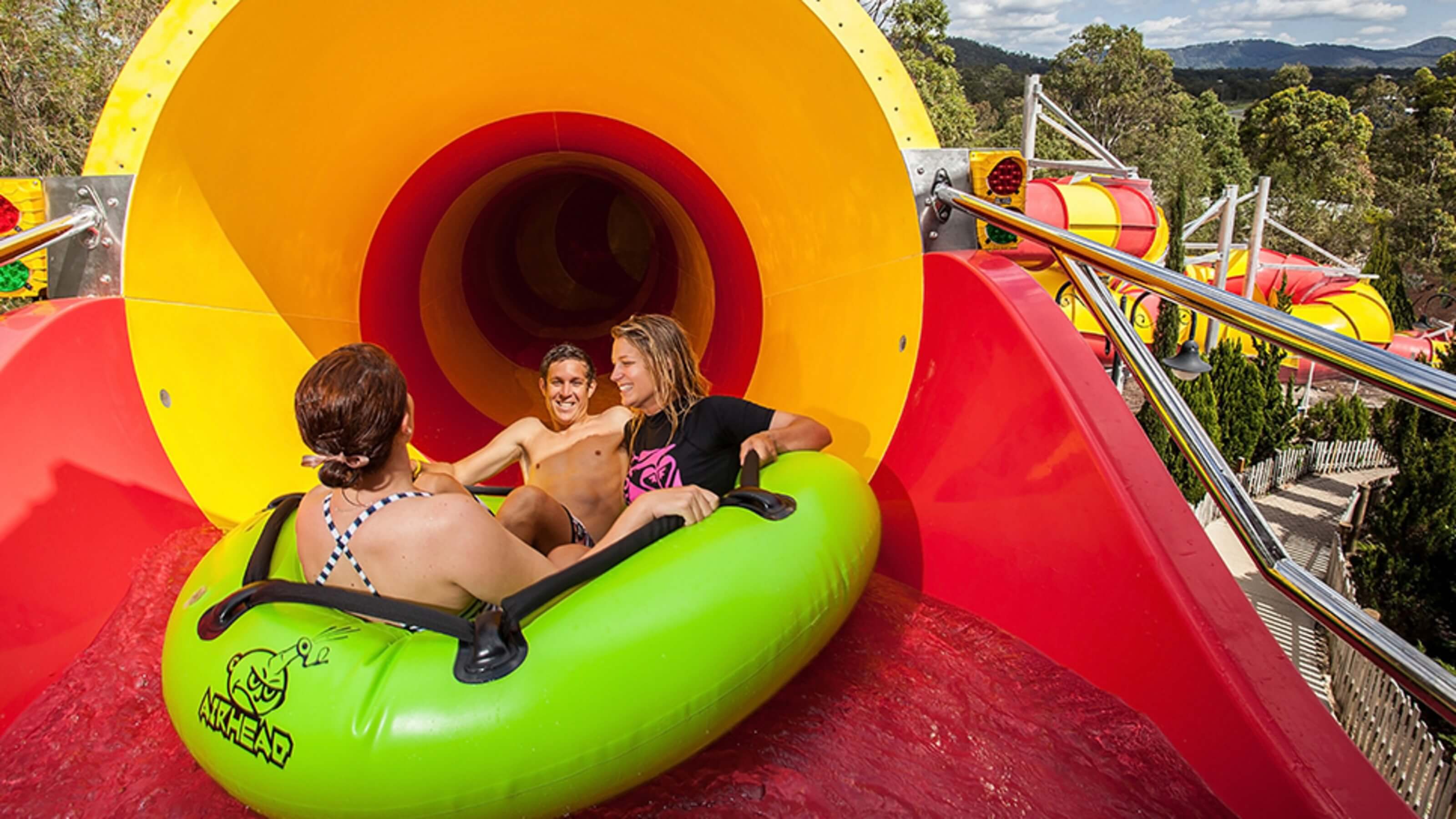 Group of riders in a green inflatable raft entering the bright red and yellow Constrictor water slide tunnel at Wet’n’Wild.