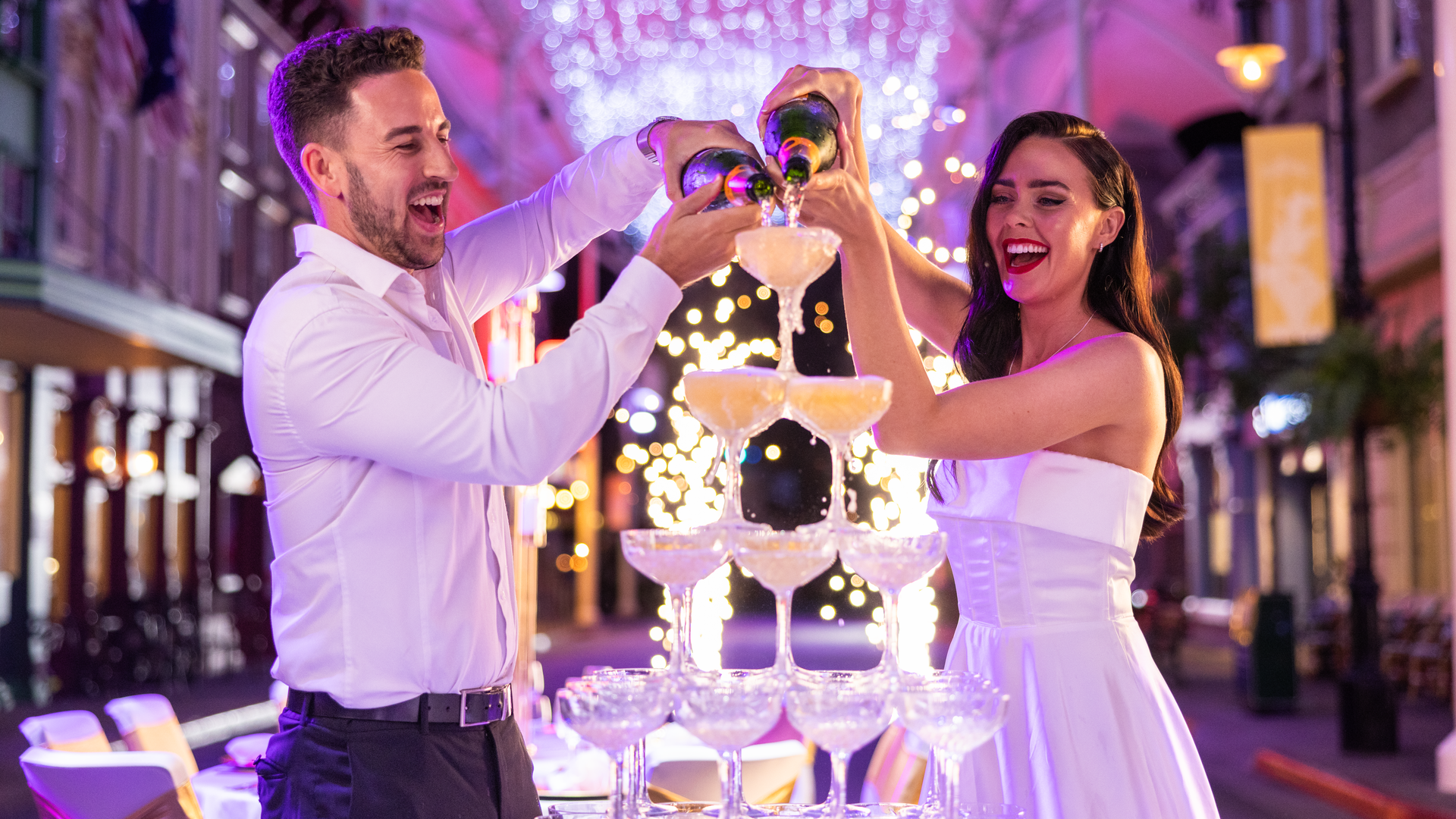 A joyful couple in formal attire pours champagne into a pyramid of glasses at an outdoor celebration, with sparkling lights and fireworks in the background.