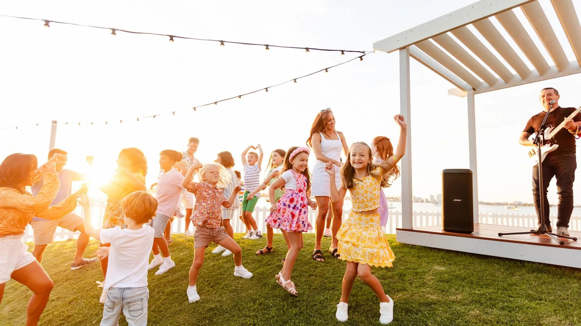 Children dancing on grass near a wooden gazebo with a musician playing guitar. Adults watch under string lights at sunset.