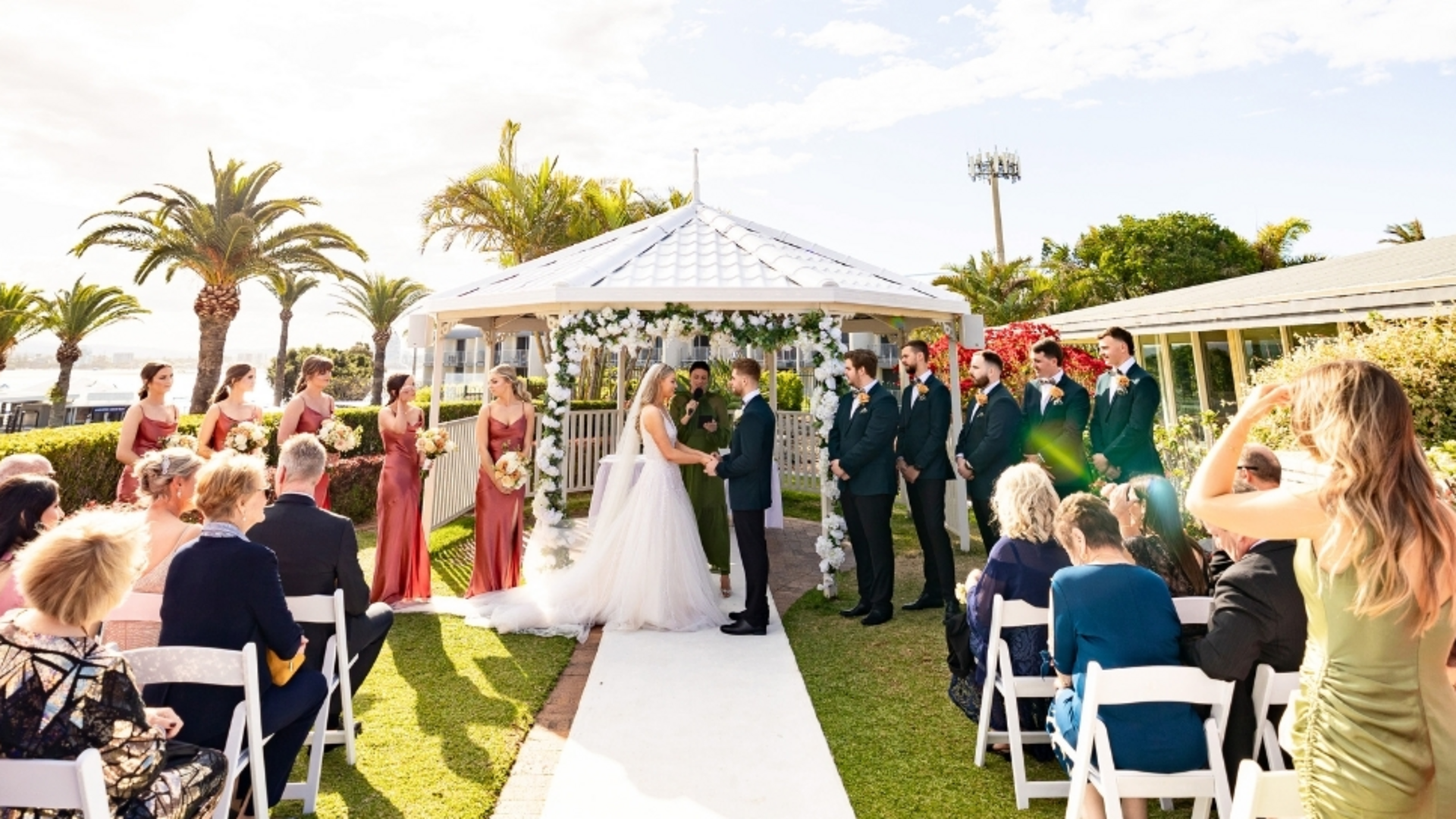 A bride and groom stand under a white gazebo, holding hands during an outdoor wedding ceremony, surrounded by bridesmaids in red dresses, groomsmen in dark suits, and seated guests under sunny skies with palm trees.