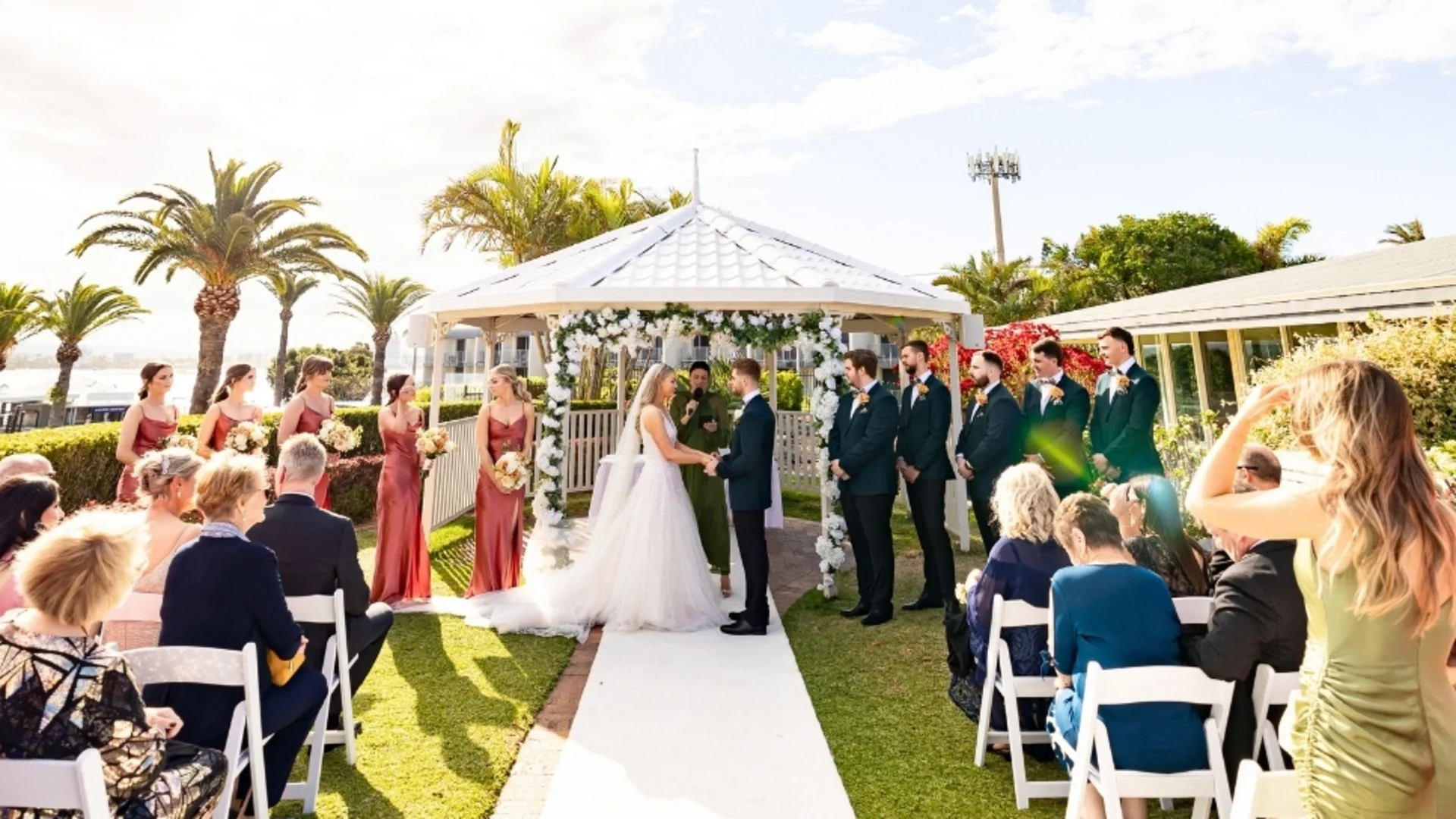 A bride and groom stand under a white gazebo, holding hands during an outdoor wedding ceremony, surrounded by bridesmaids in red dresses, groomsmen in dark suits, and seated guests under sunny skies with palm trees.