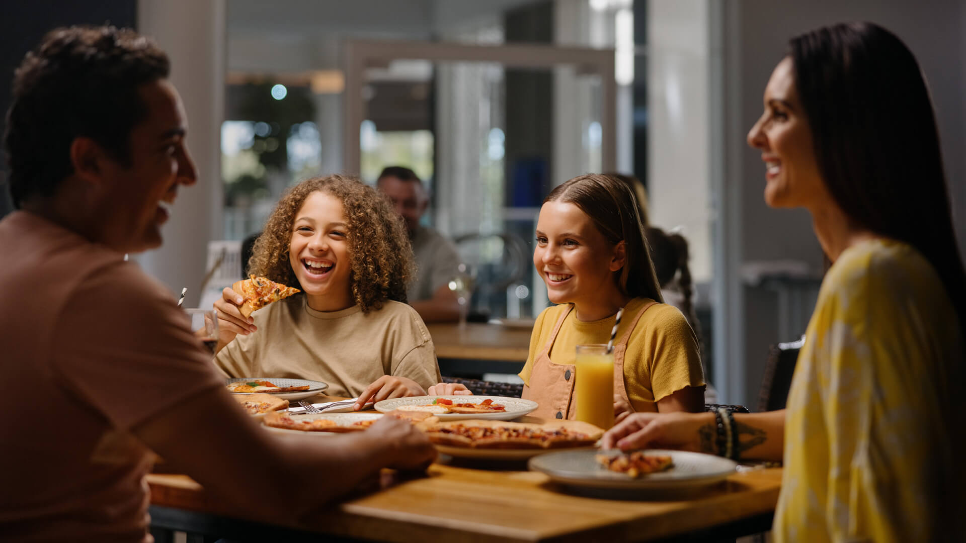 A family of four happily eats pizza at a dining table in Melrose Italian Restaurant at Sea World Resort. The two children are smiling, and the table is filled with pizza and drinks.