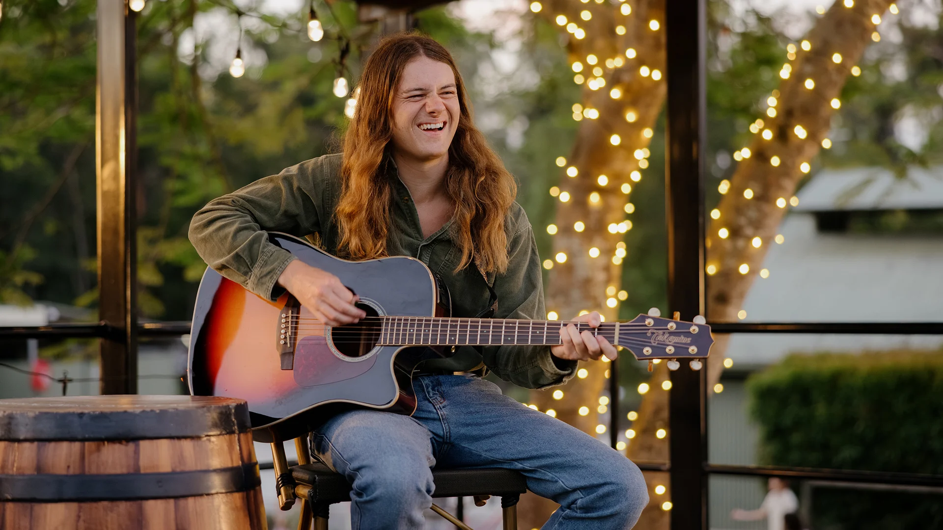 A smiling person with long hair sits on a stool, playing an acoustic guitar outdoors. They wear a green jacket and jeans, with string lights and trees in the background.