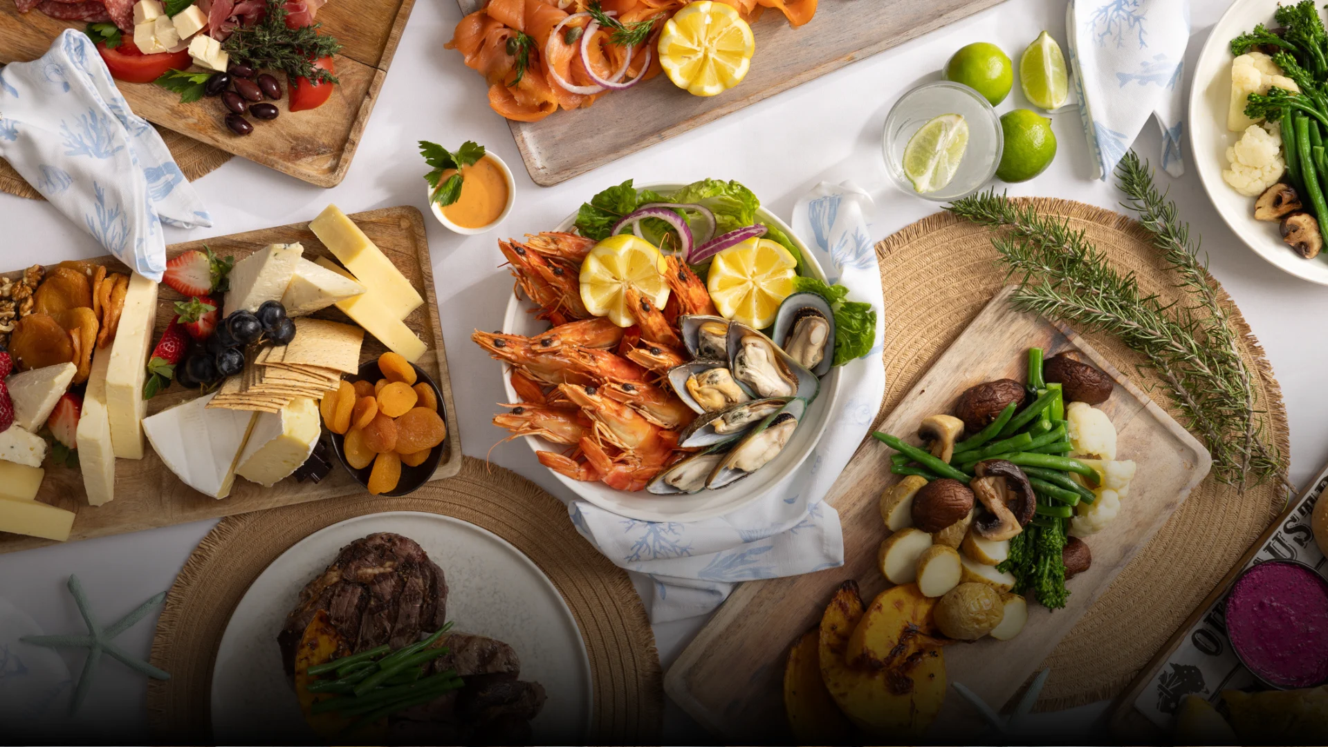 A table set with assorted dishes including grilled shrimp, mussels, sliced smoked salmon, cheese and fruit platter, steak with vegetables, and a plate of green beans, potatoes, and mushrooms, alongside limes and a drink.