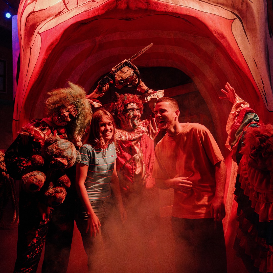 Two people pose and smile with actors in monster costumes in a foggy, eerie setting with a giant mouth entrance and colorful lights, creating a spooky and festive atmosphere.