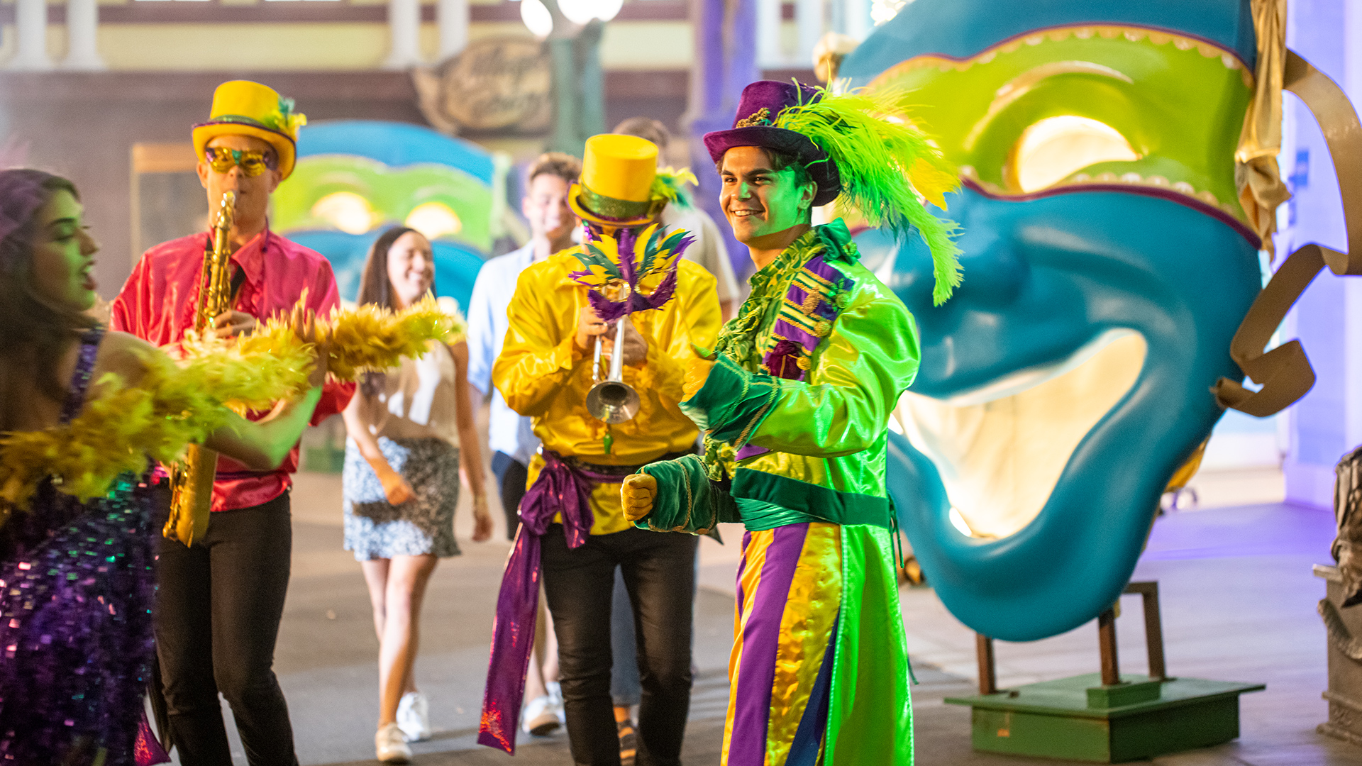 People in colorful Mardi Gras costumes and hats play instruments and dance in front of large decorative masks, creating a festive parade atmosphere.