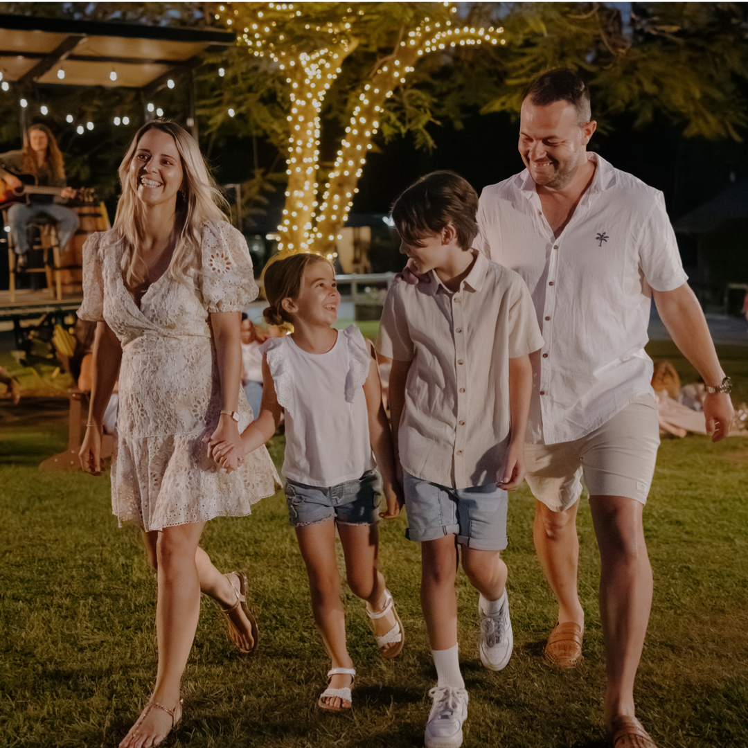 A smiling family of four, two adults and two children, walk hand in hand on a grassy area at night, with string lights wrapped around a tree in the background.