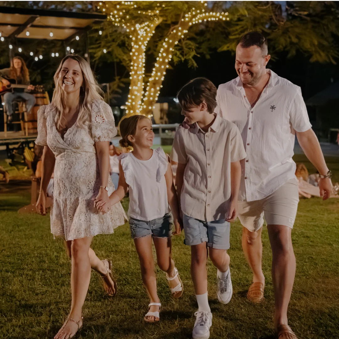 A smiling family of four, two adults and two children, walk hand in hand on a grassy area at night, with string lights wrapped around a tree in the background.