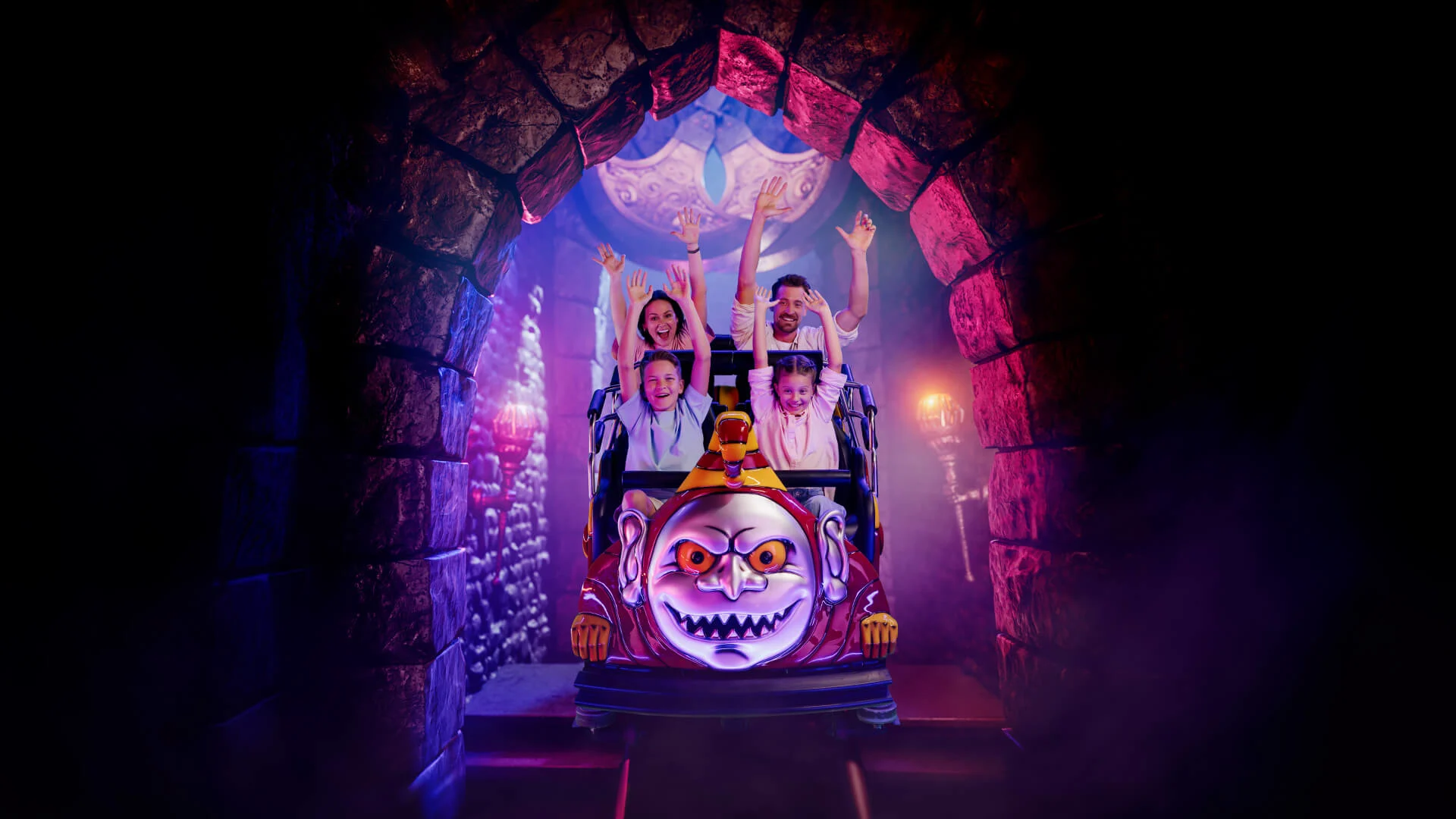 Four people ride a spooky-themed roller coaster car with a scary face design, raising their arms as they exit a dark, stone tunnel lit with purple light.