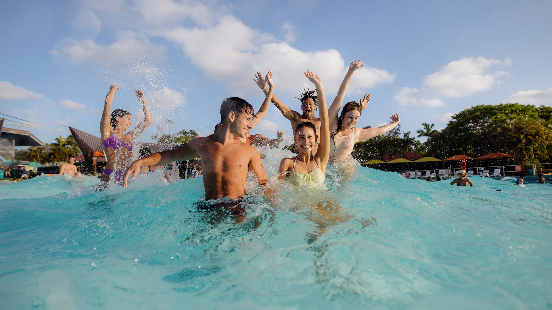 A group of young people in swimwear enjoy splashing and playing in a wave pool on a sunny day, with clear blue water, trees, and other swimmers in the background.