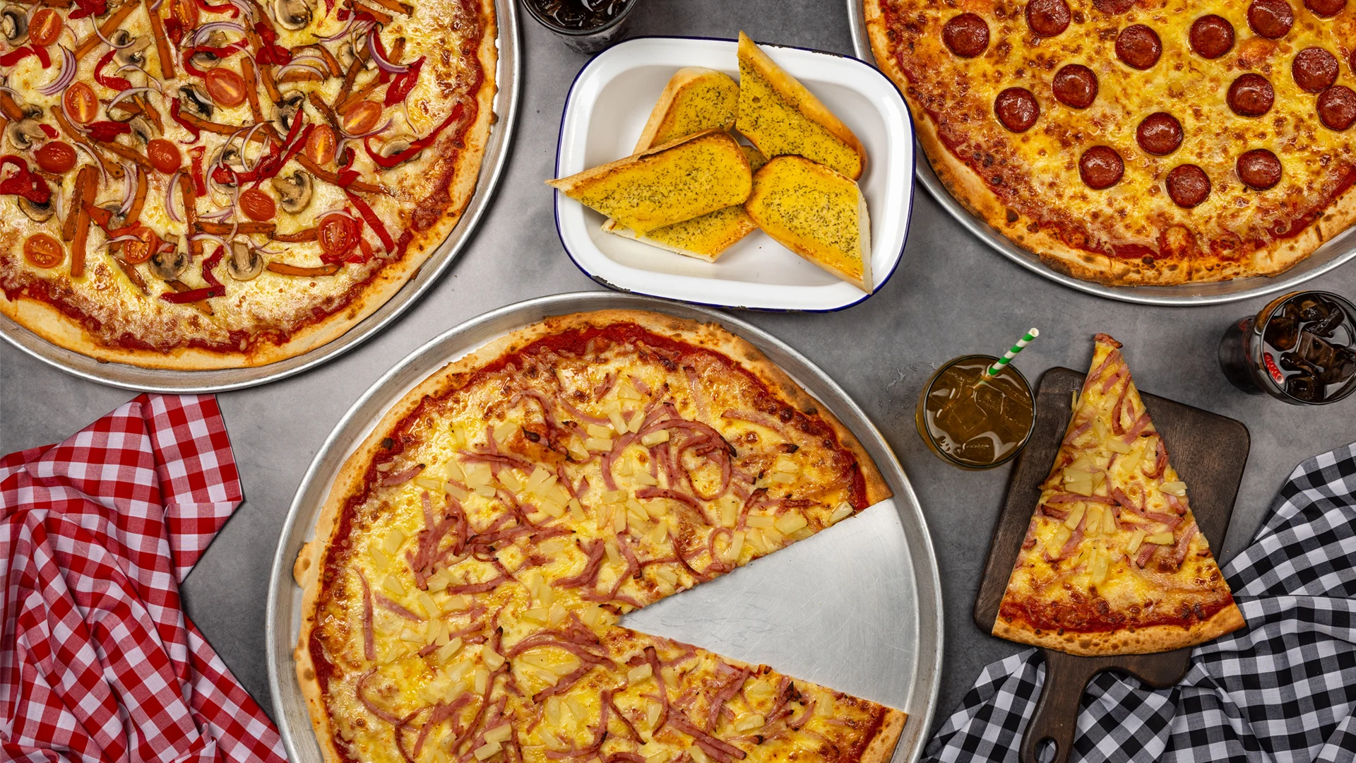 Four pizzas with different toppings, a serving of garlic bread, and several glasses of soda are arranged on a table with red and black checkered napkins. One pizza has a slice missing and placed on a wooden board.