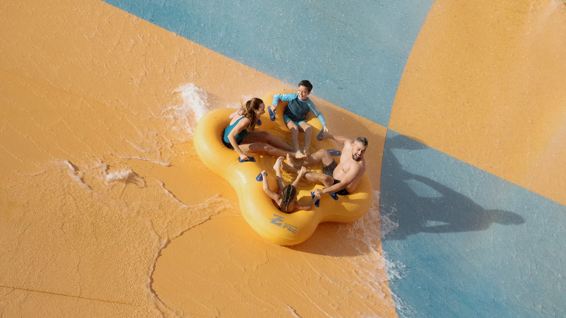 Four people ride together in a large yellow inflatable raft on a water slide, wearing swimsuits and rash guards, with water splashing around them on a colorful slide.