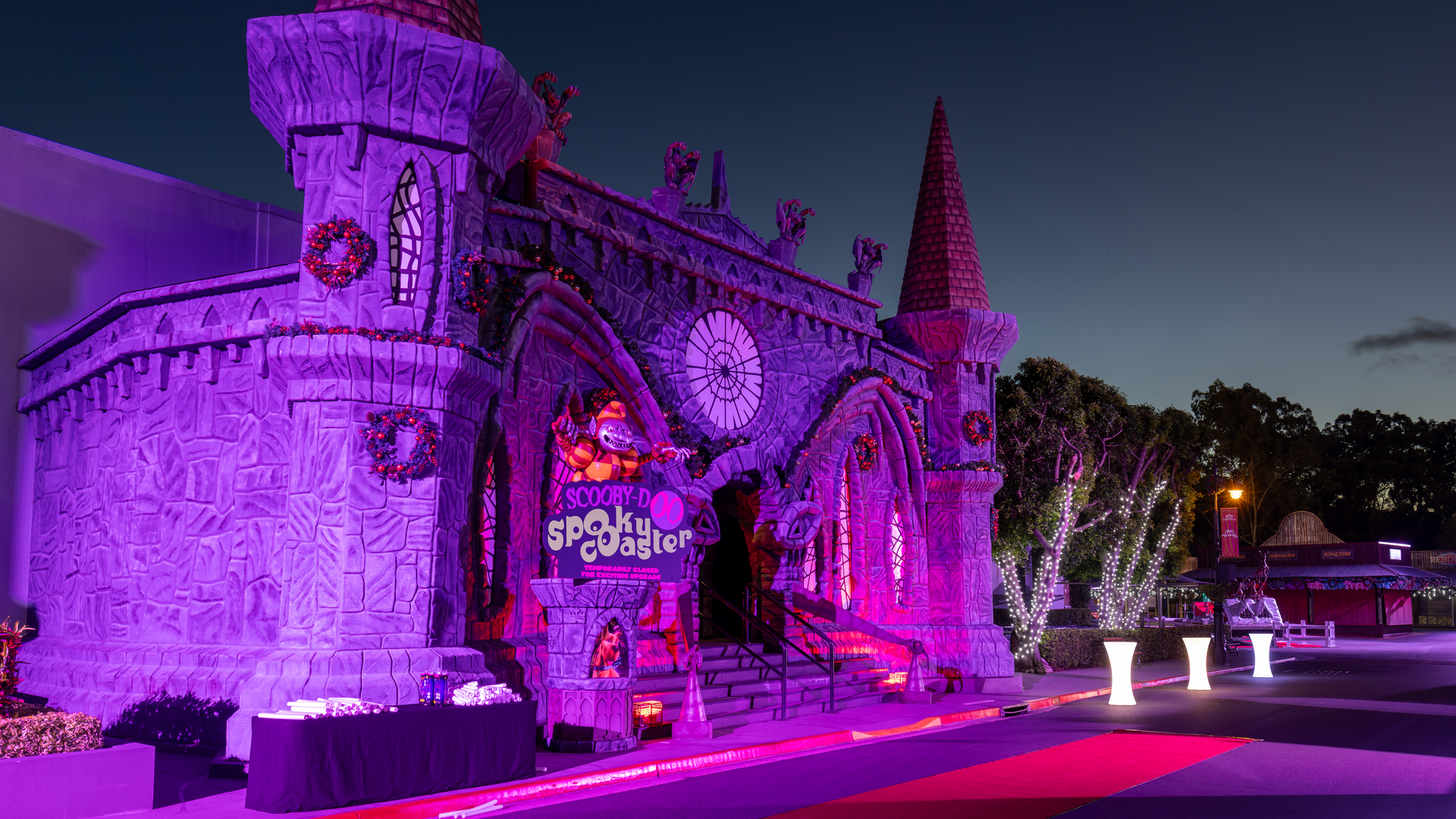 A large, castle-like building decorated with purple and orange lights at night, featuring a "Scooby Spooky Castle" sign at the entrance and a red carpet leading up to the stairs.