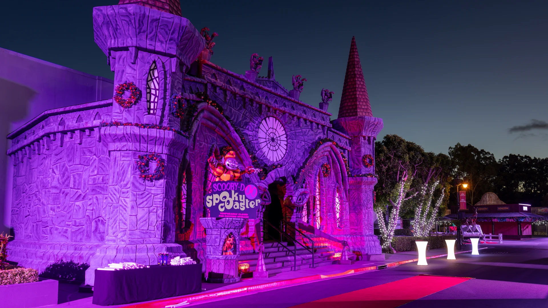 A large, castle-like building decorated with purple and orange lights at night, featuring a "Scooby Spooky Castle" sign at the entrance and a red carpet leading up to the stairs.