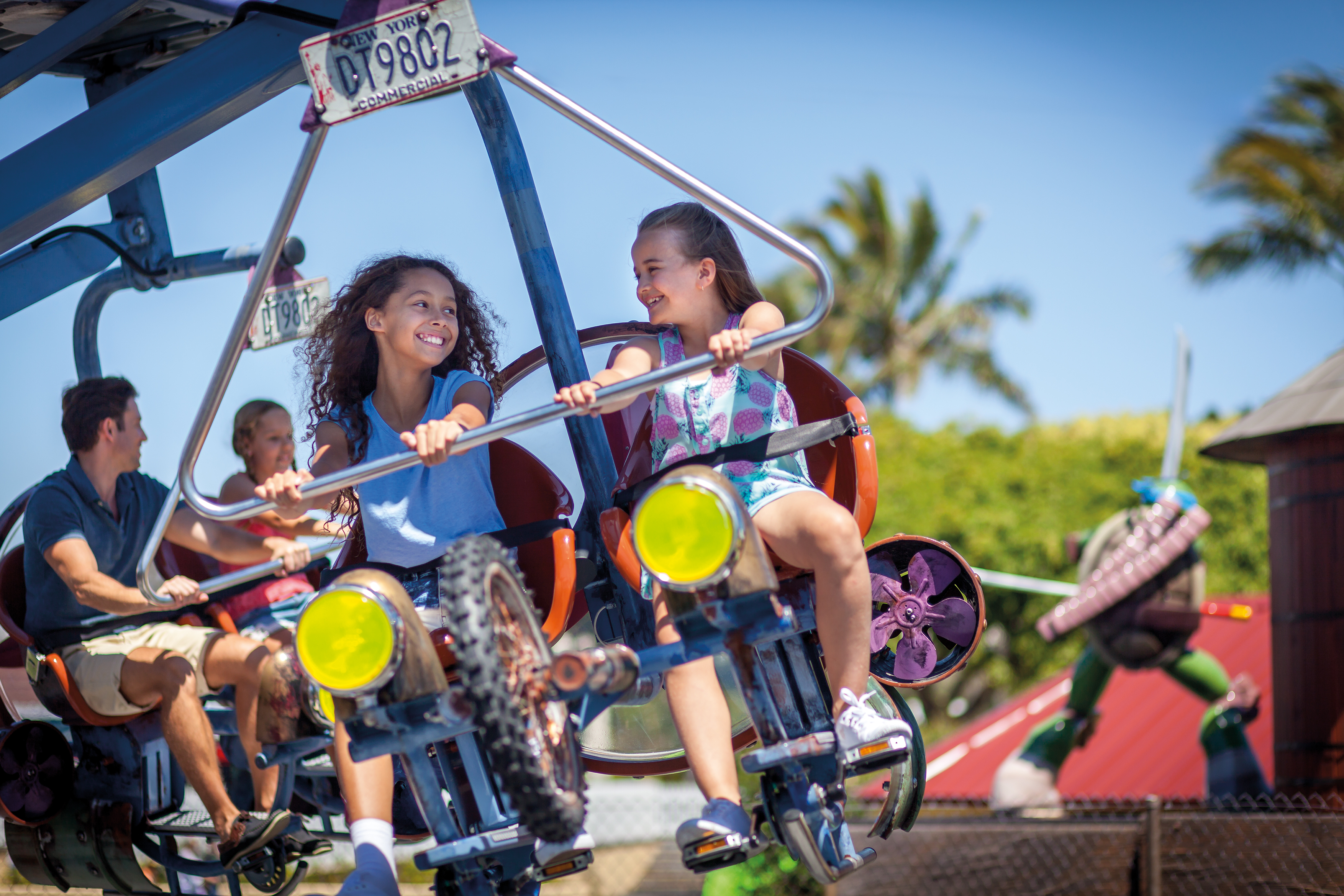 Two girls smiling and riding a pedal-powered amusement park ride, with other riders and palm trees visible in the background.