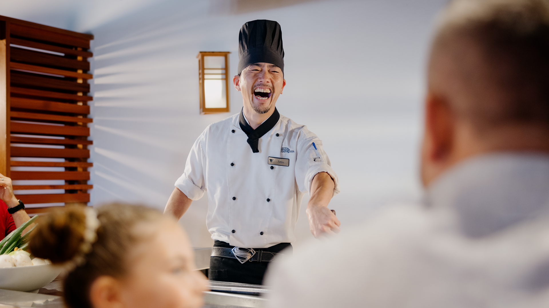 A chef in a white uniform and black hat laughs joyfully while interacting with guests seated at a counter in a brightly lit restaurant.