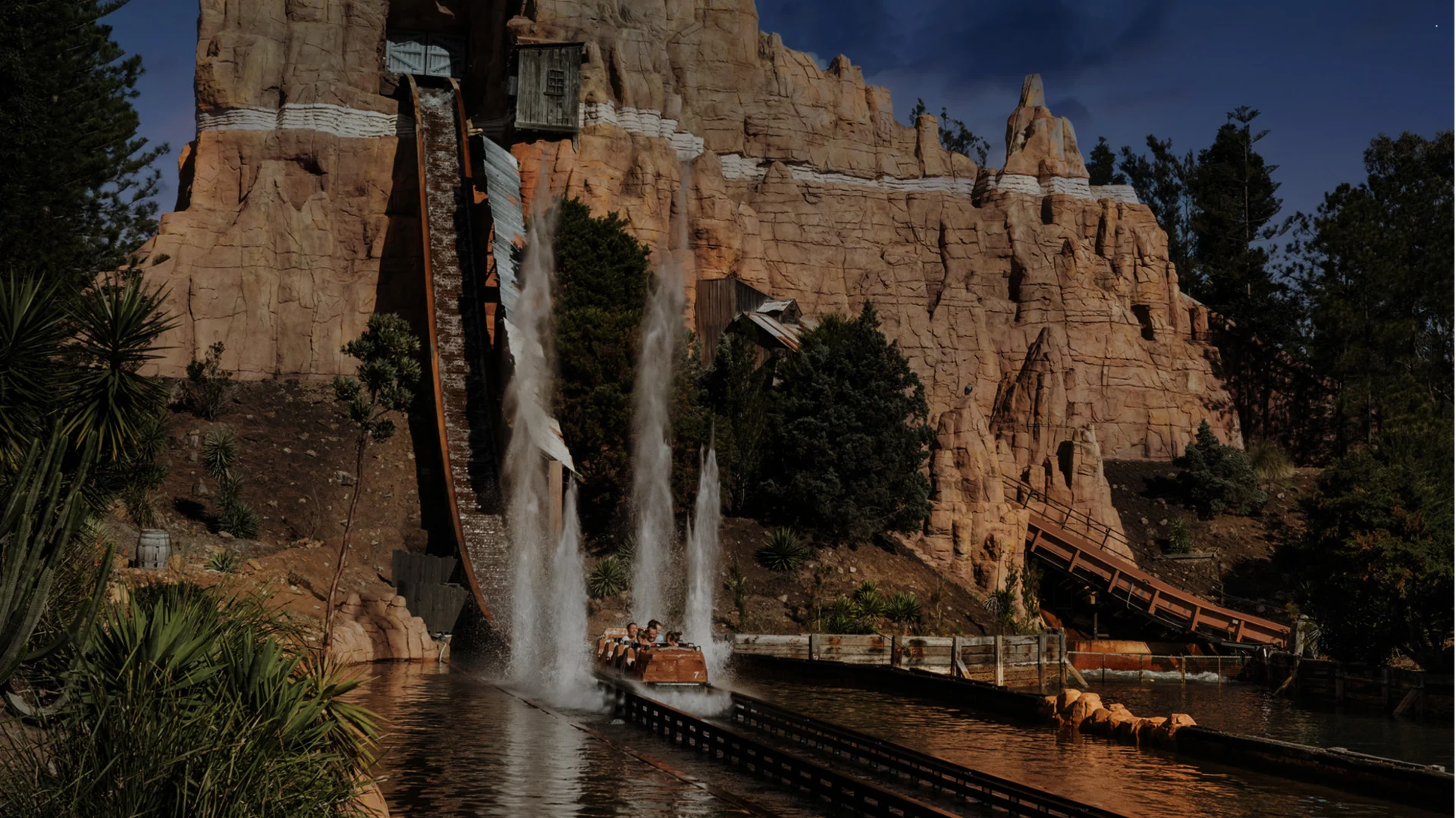 A log flume ride splashes down a steep drop, sending water spraying into the air in front of a rocky, mountain-themed amusement park attraction with trees and greenery surrounding the ride.