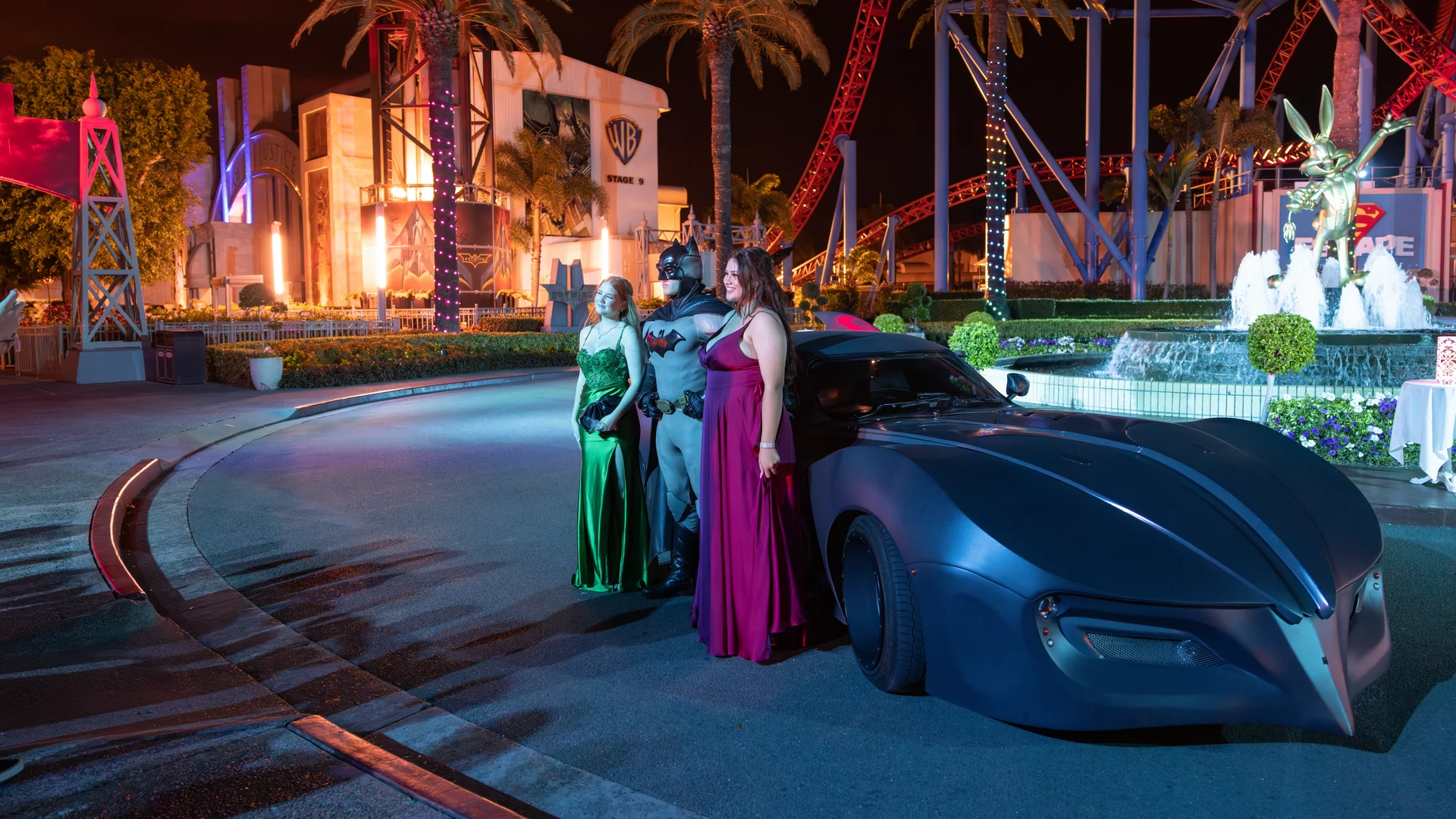 Three people in formal gowns pose with a person dressed as Batman in front of a Batmobile replica at a theme park, with palm trees, roller coaster tracks, and illuminated buildings in the background at night.