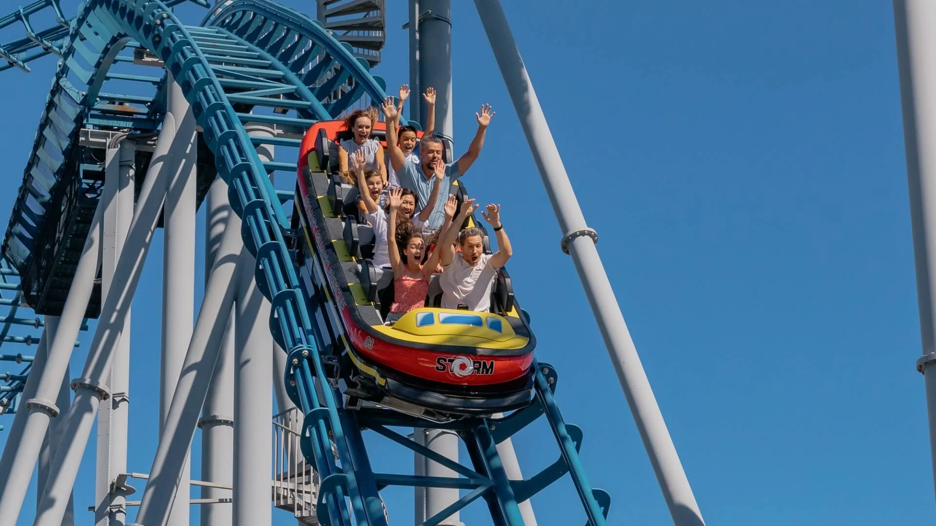 People on a roller coaster ride, Storm Coaster at Sea World, with raised arms, descending a steep track under a clear blue sky.