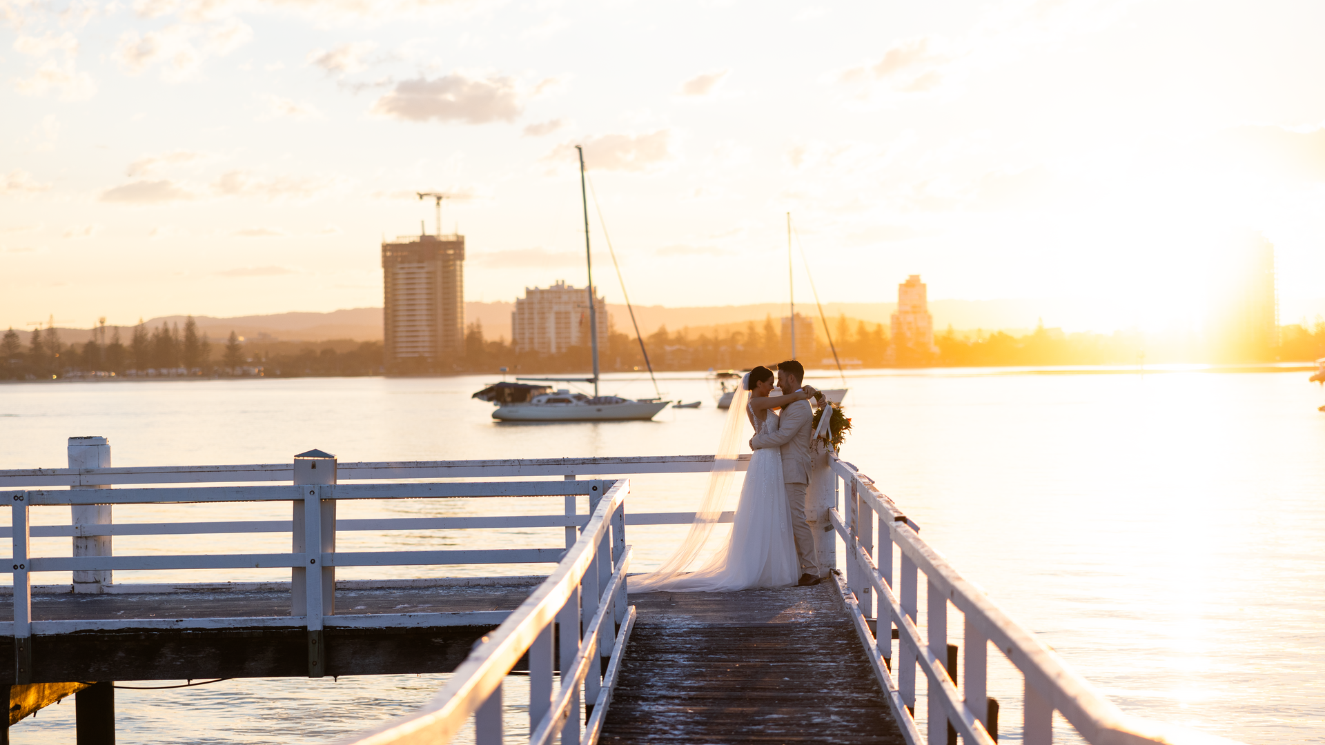 A bride and groom embrace on a wooden pier at sunset, with calm water, sailboats, and city buildings in the background. The golden sunlight creates a warm, romantic atmosphere.