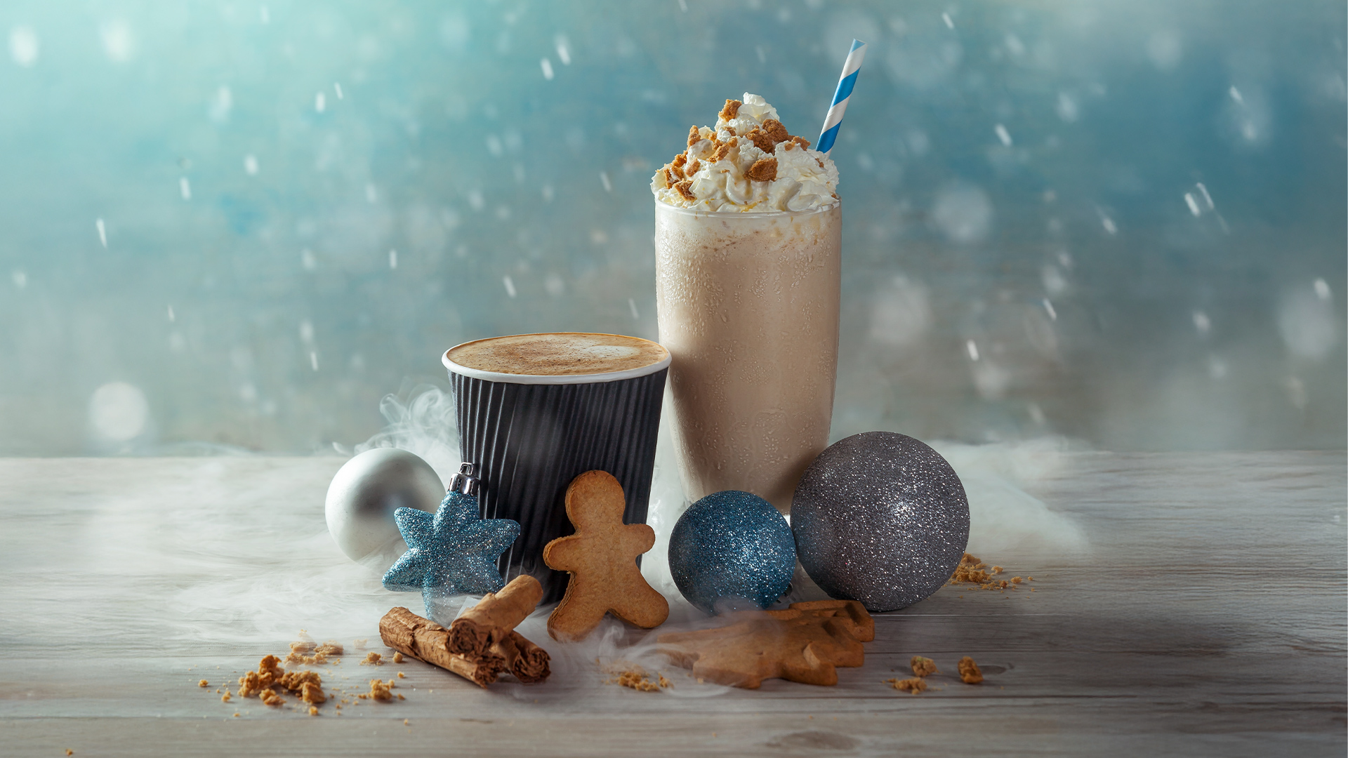 A festive scene with a latte in a black cup, a milkshake topped with whipped cream and caramel, gingerbread cookies, silver and blue Christmas ornaments, and snow-like decoration on a wooden surface.