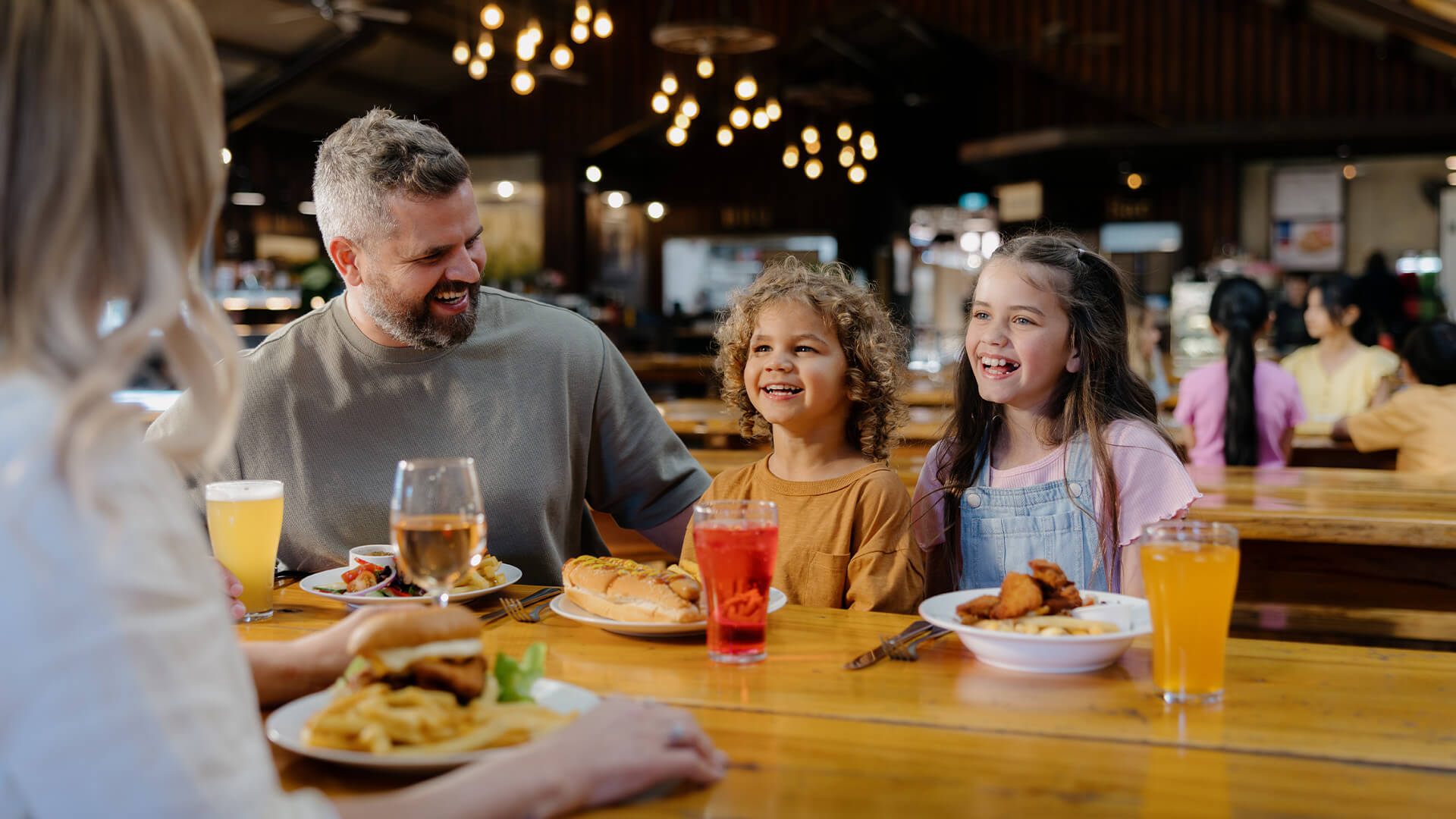 A family of four sits at a restaurant table, smiling and laughing together. They have plates of food and drinks in front of them, enjoying a cheerful meal in a warmly lit, casual dining setting.