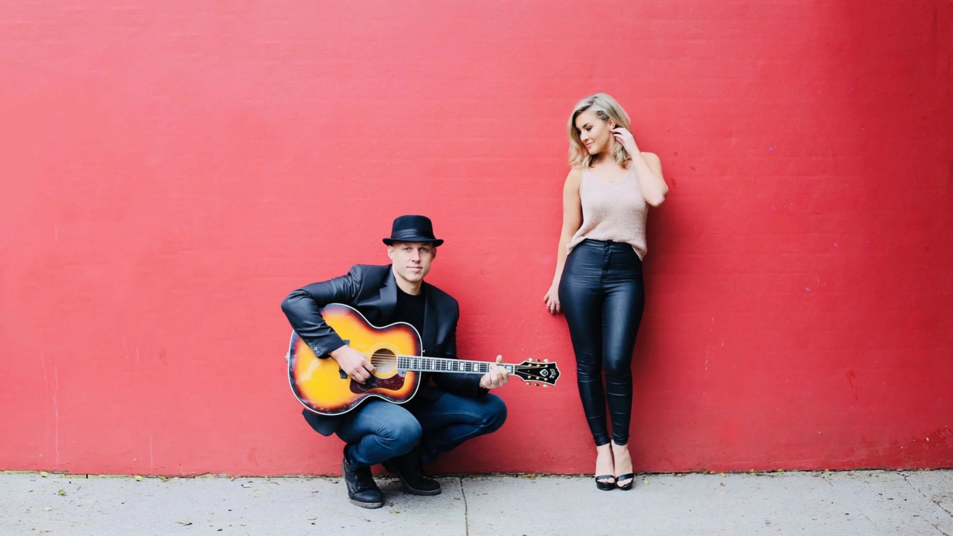 A man in a hat kneels playing an acoustic guitar next to a woman standing against a bright red wall. The woman is looking down and touching her hair, while the man looks toward the camera.