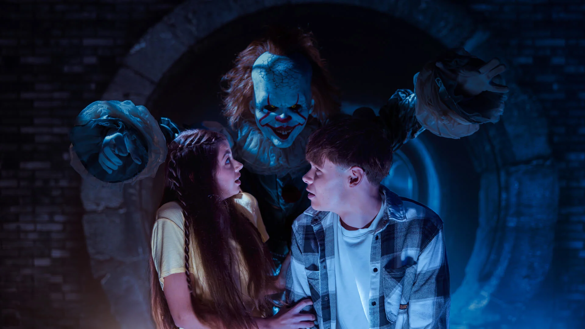 Two frightened teenagers stand in front of a dark tunnel while a sinister clown (Pennywise) with orange hair and white makeup looms menacingly behind them, ready to attack.