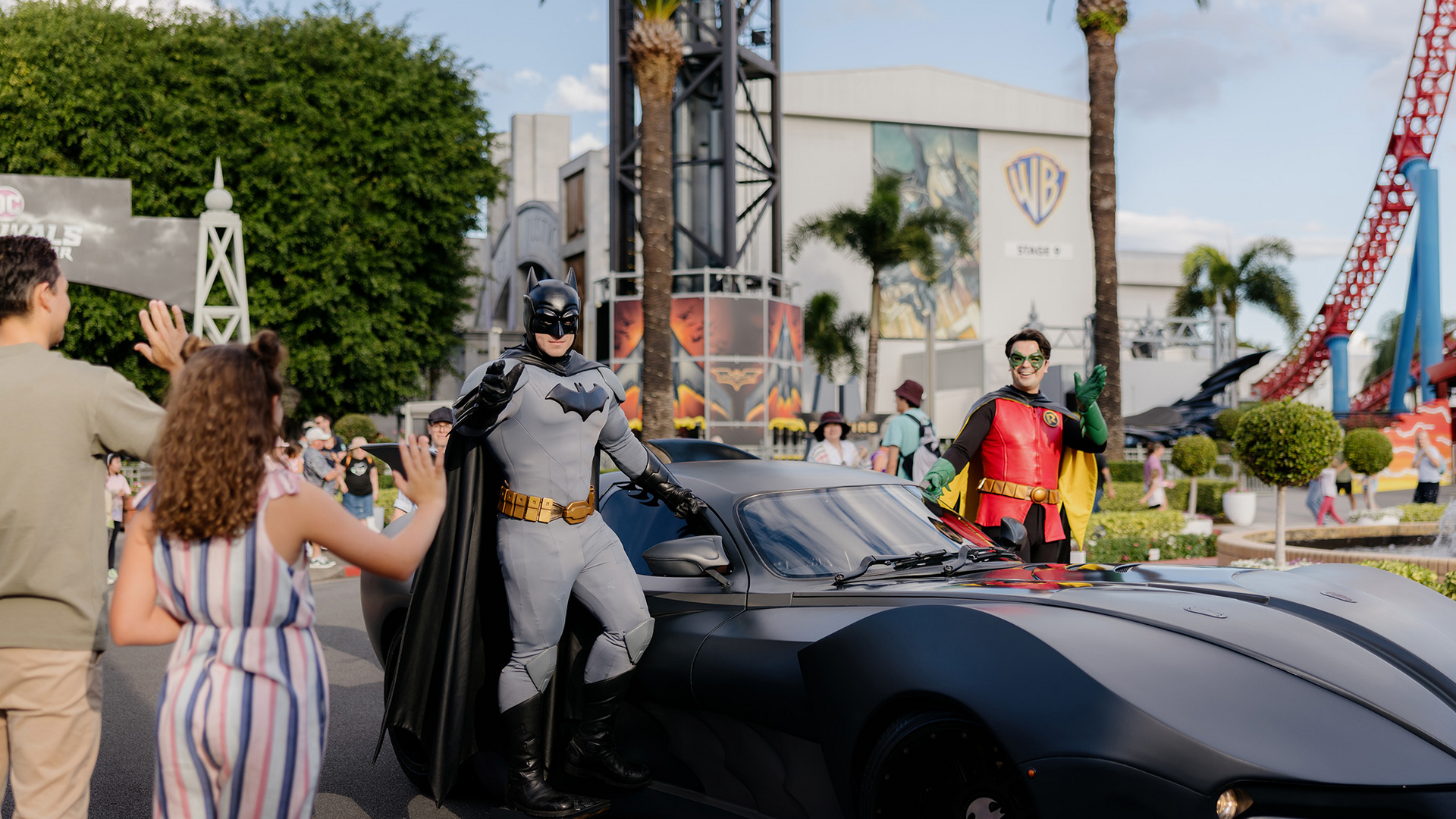 People interact with costumed Batman and Robin characters standing by a sleek black Batmobile at an outdoor theme park, with palm trees, colorful buildings, and a red roller coaster in the background.