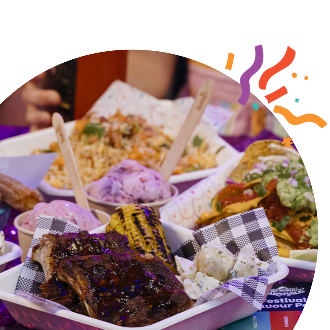 A colorful assortment of food on a table, including churros, ribs, grilled corn, rice, and ice cream, with people in the background. The scene is festive with confetti-like decorations.