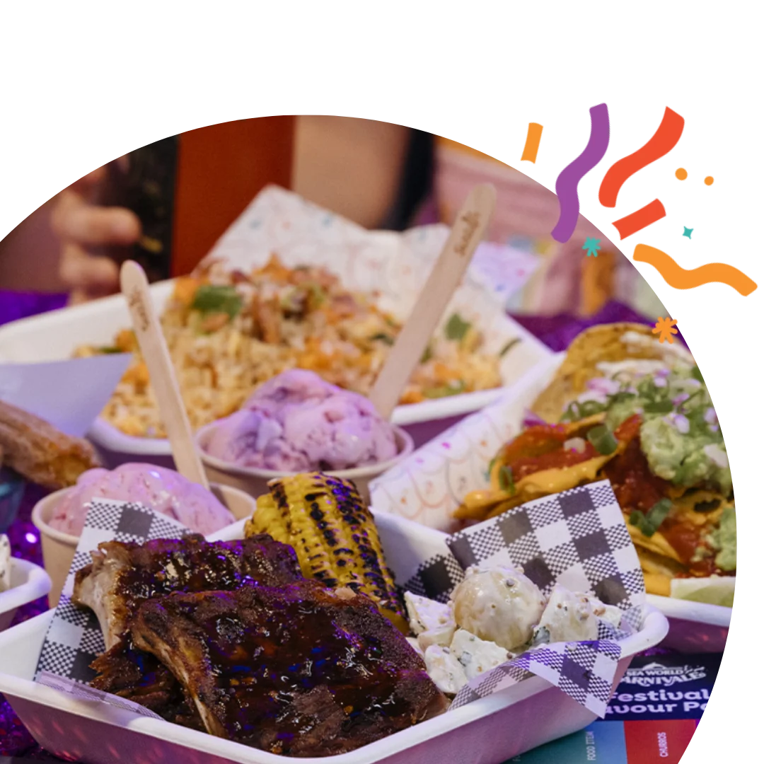 A colorful assortment of food on a table, including churros, ribs, grilled corn, rice, and ice cream, with people in the background. The scene is festive with confetti-like decorations.