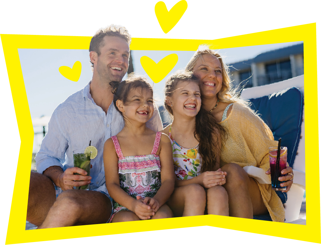 A smiling family of four sits close together outdoors, with two young girls in the middle and parents on either side, each holding drinks. Yellow cartoon hearts and a border decorate the cheerful scene.