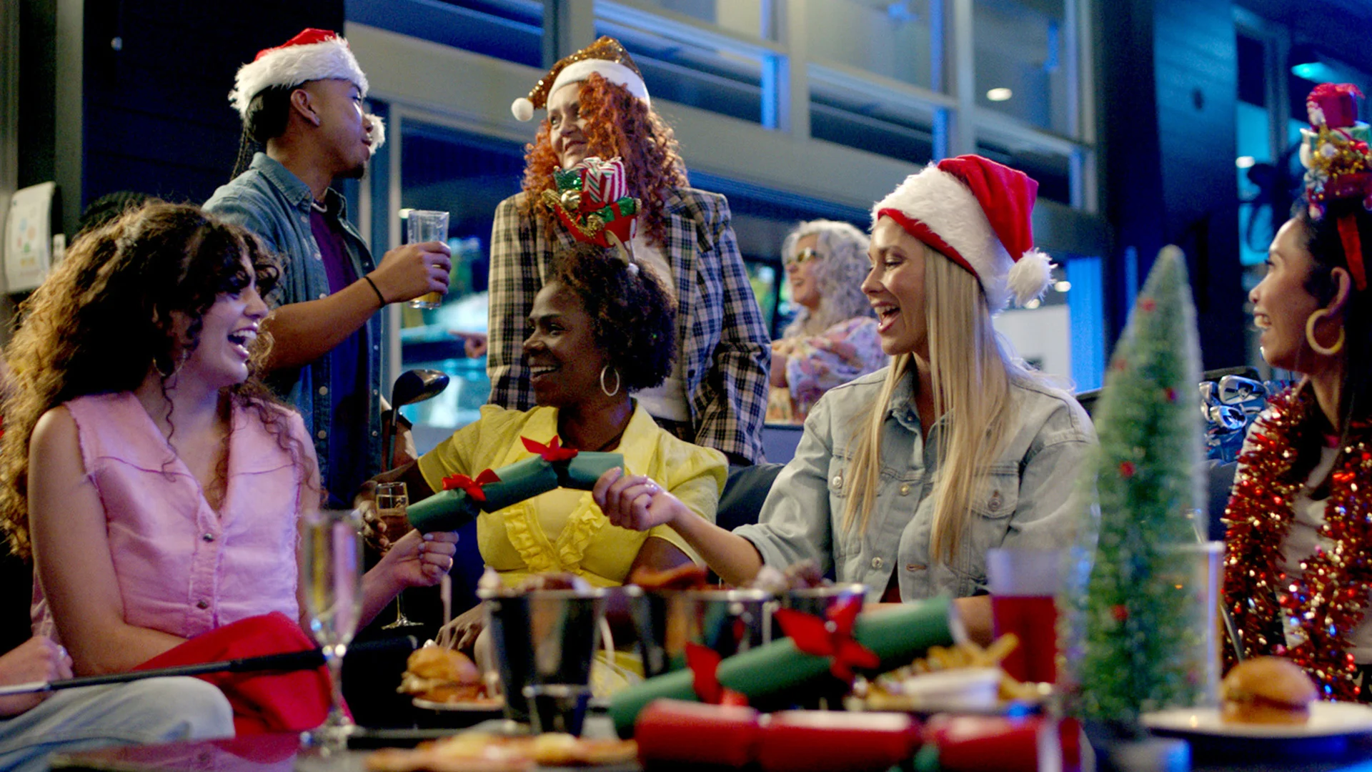 A group of people in festive hats laugh and celebrate at a holiday party, pulling Christmas crackers at a decorated table with food and drinks.
