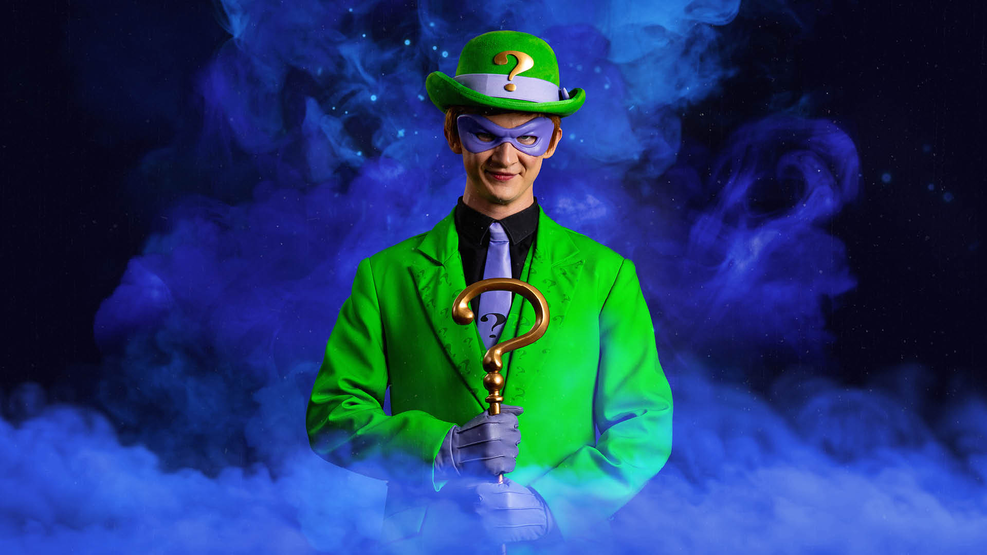 A person in a green suit, green hat with a question mark, and purple mask holds a gold cane shaped like a question mark, with green smoke in the background.
