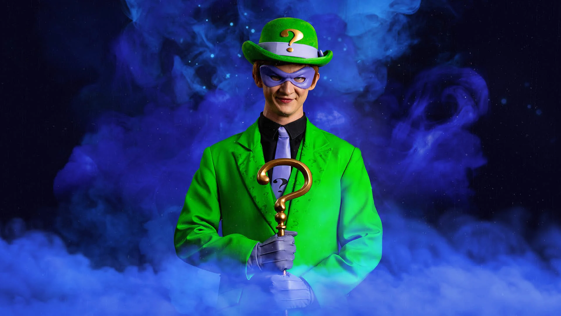 A person in a green suit, green hat with a question mark, and purple mask holds a gold cane shaped like a question mark, with green smoke in the background.