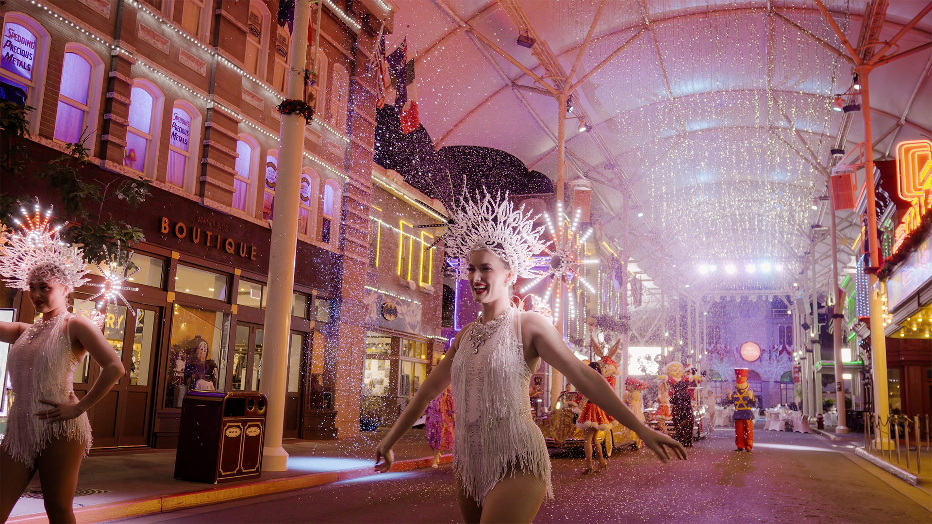 Two performers in sparkling white costumes and spiky headdresses dance under festive lights and falling confetti on a lively, colorful street with decorated buildings and an excited crowd.