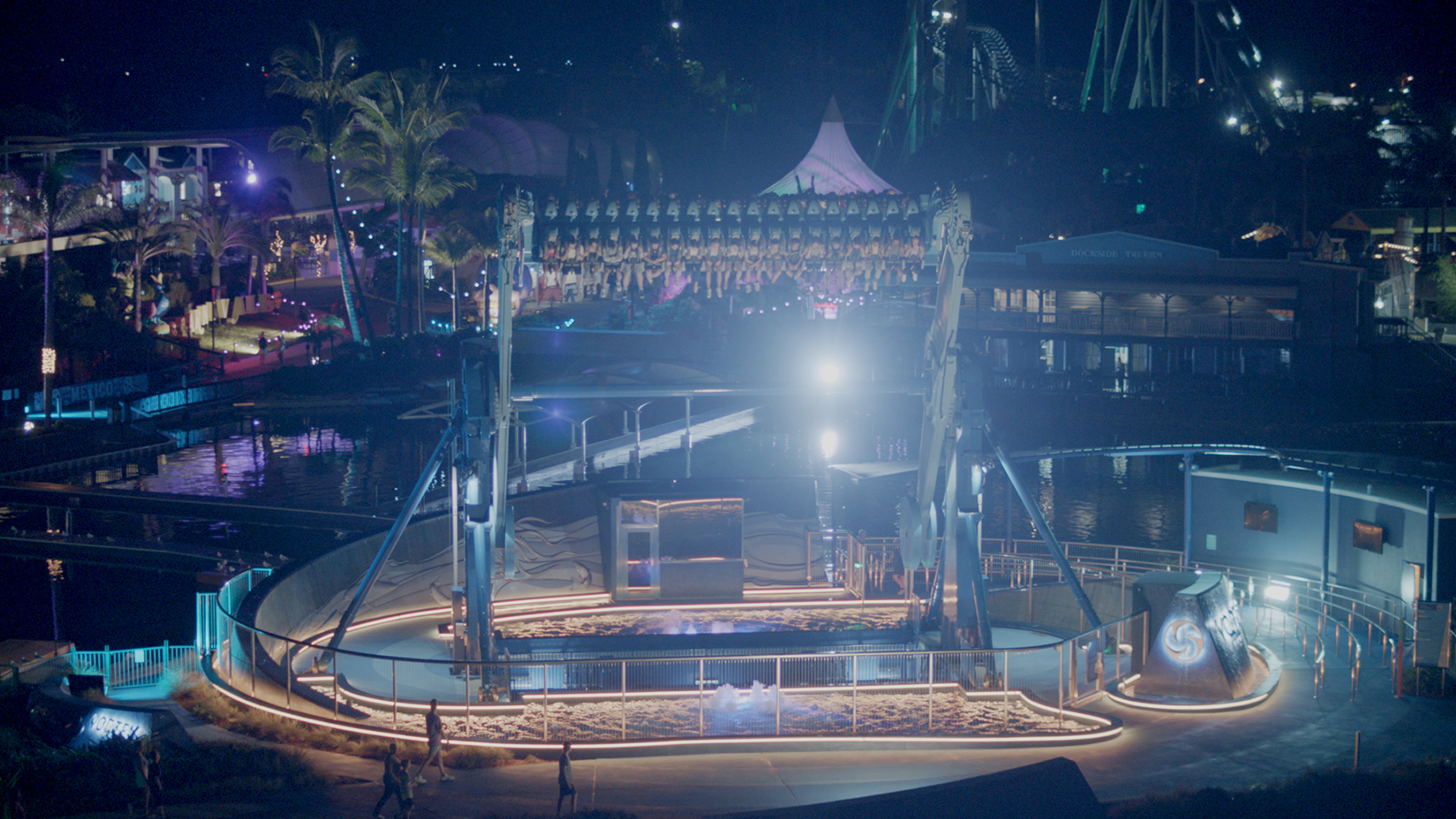 An amusement park ride spins riders upside down at night, illuminated by bright lights. The surrounding park areas and attractions are also visible, with paths winding through the scene.