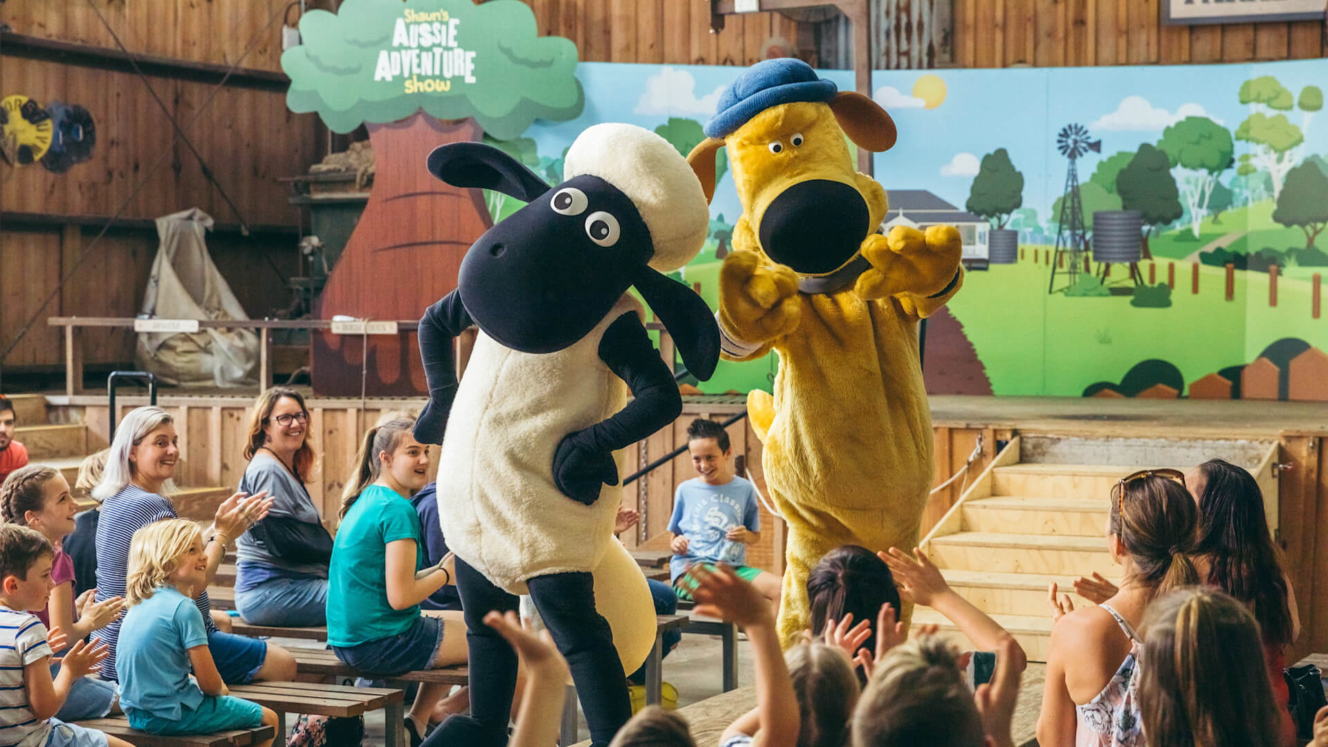 Two people in Shaun the Sheep and Bitzer dog costumes perform on stage in front of an audience of children and adults at an indoor venue.
