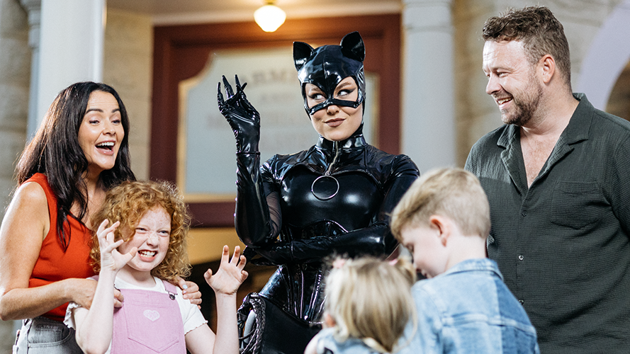 A woman in a Catwoman costume poses with a smiling man, woman, and children. The kids make playful claw gestures, and everyone appears to be enjoying themselves indoors.