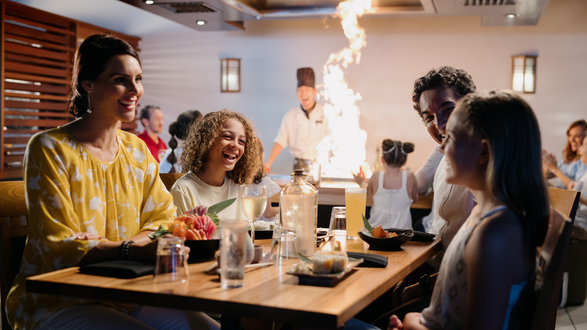 A family sits at a restaurant table with a chef in the background, performing a teppanyaki-style cooking display with flames. They are smiling and appear to be enjoying the experience at Hatsuhana Japanese Restaurant.