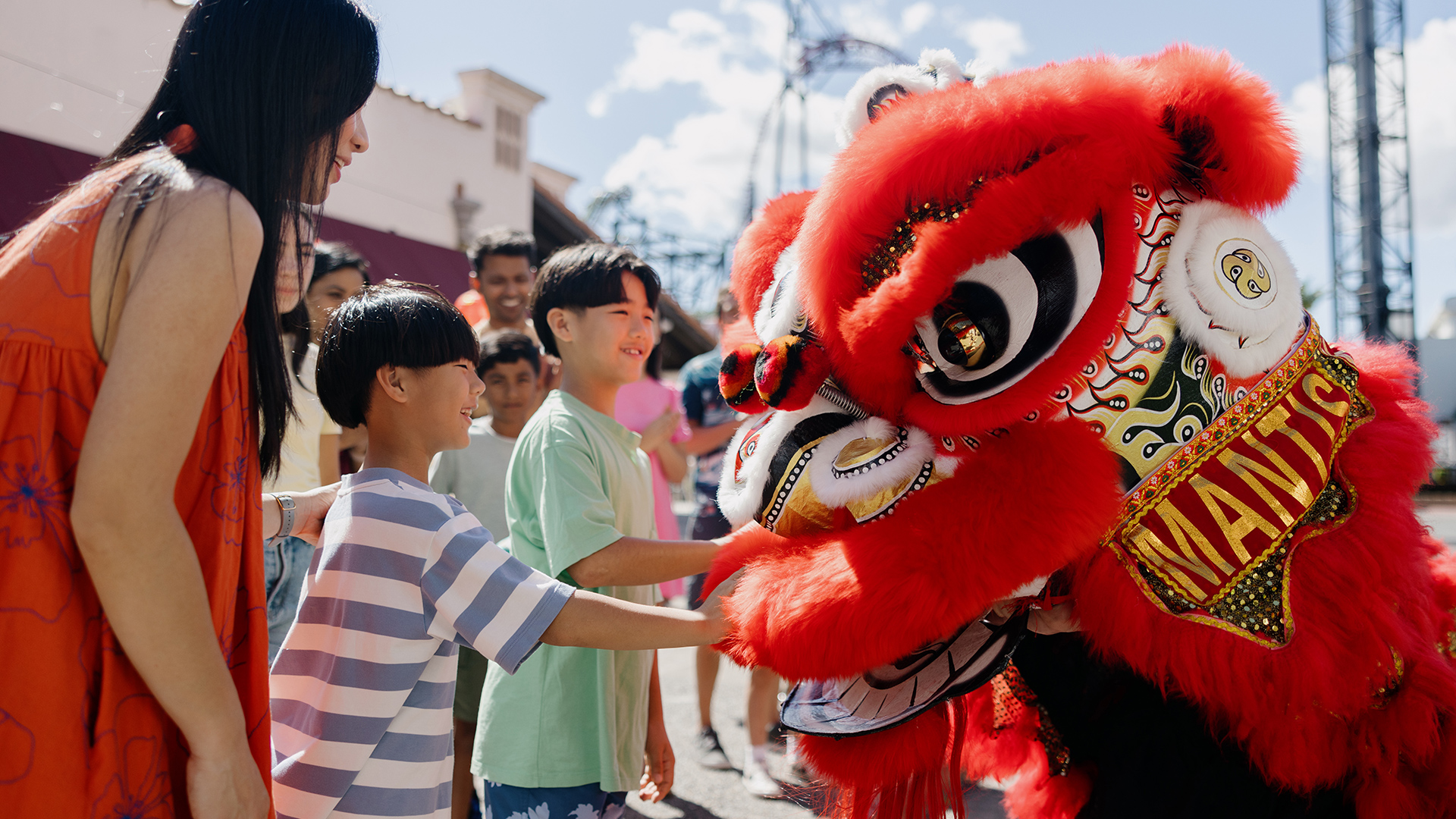 Children and adults smile and reach out to touch a vibrant red lion costume during a festive outdoor lion dance performance under a sunny sky. The atmosphere is lively and joyful.