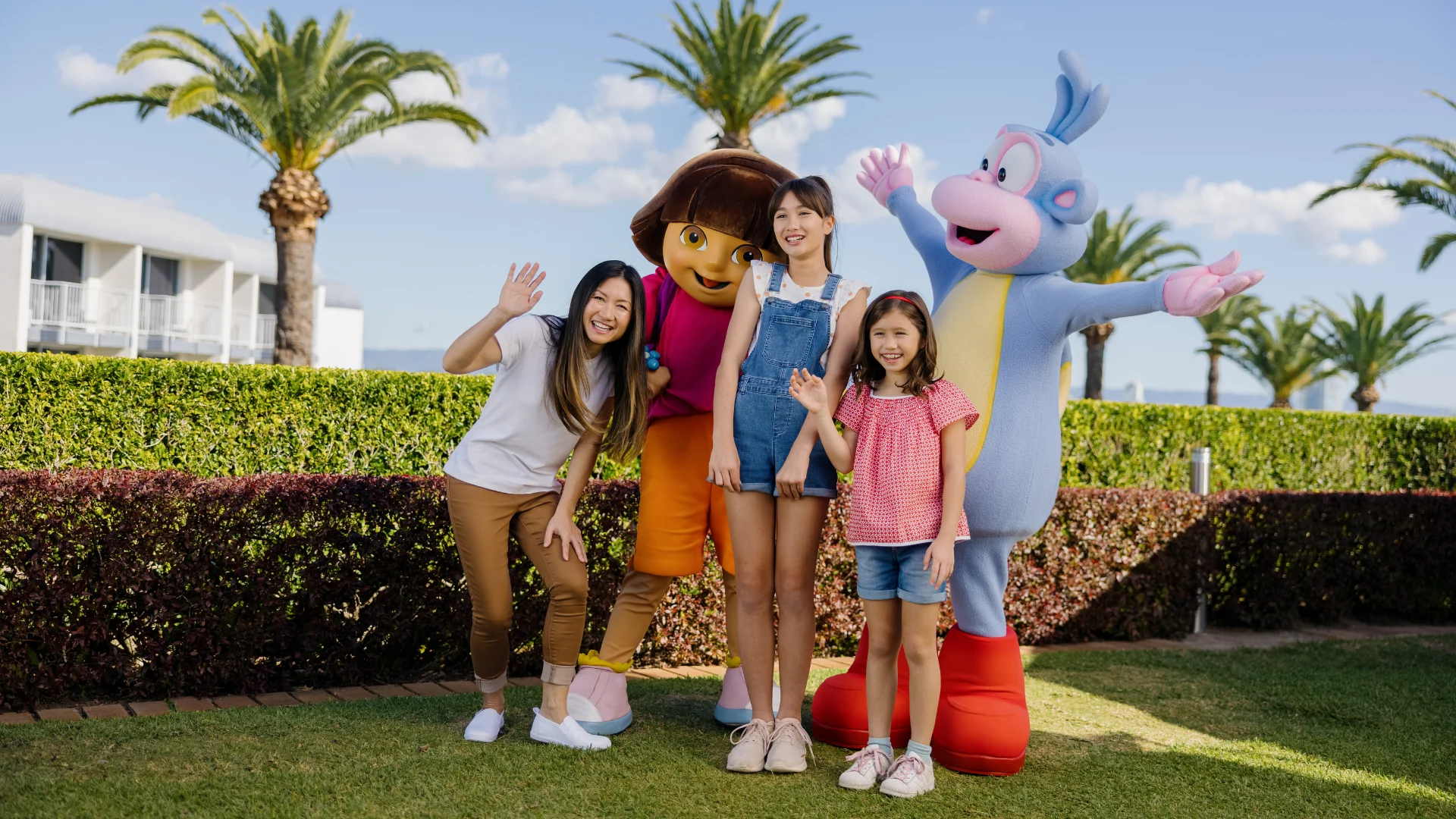 Three people, two adults and one child, smile and wave while posing outdoors with two costumed cartoon characters in front of palm trees and a hedge on a sunny day.