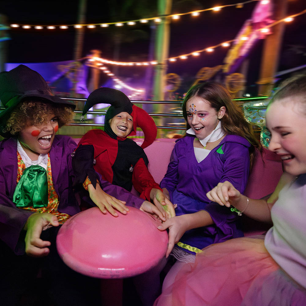 Four children in colorful costumes laugh and spin on a pink amusement park ride at night, surrounded by string lights and festive decorations.