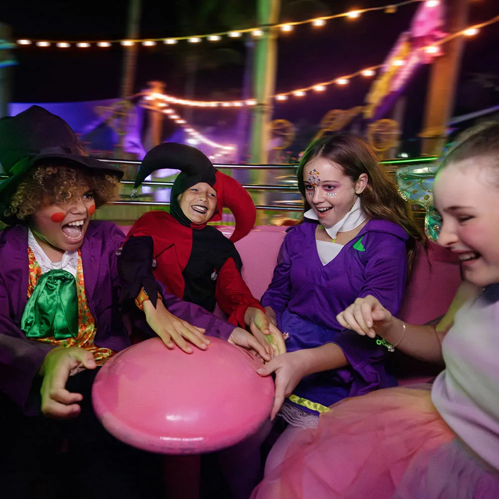 Four children in colorful costumes laugh and spin on a pink amusement park ride at night, surrounded by string lights and festive decorations.