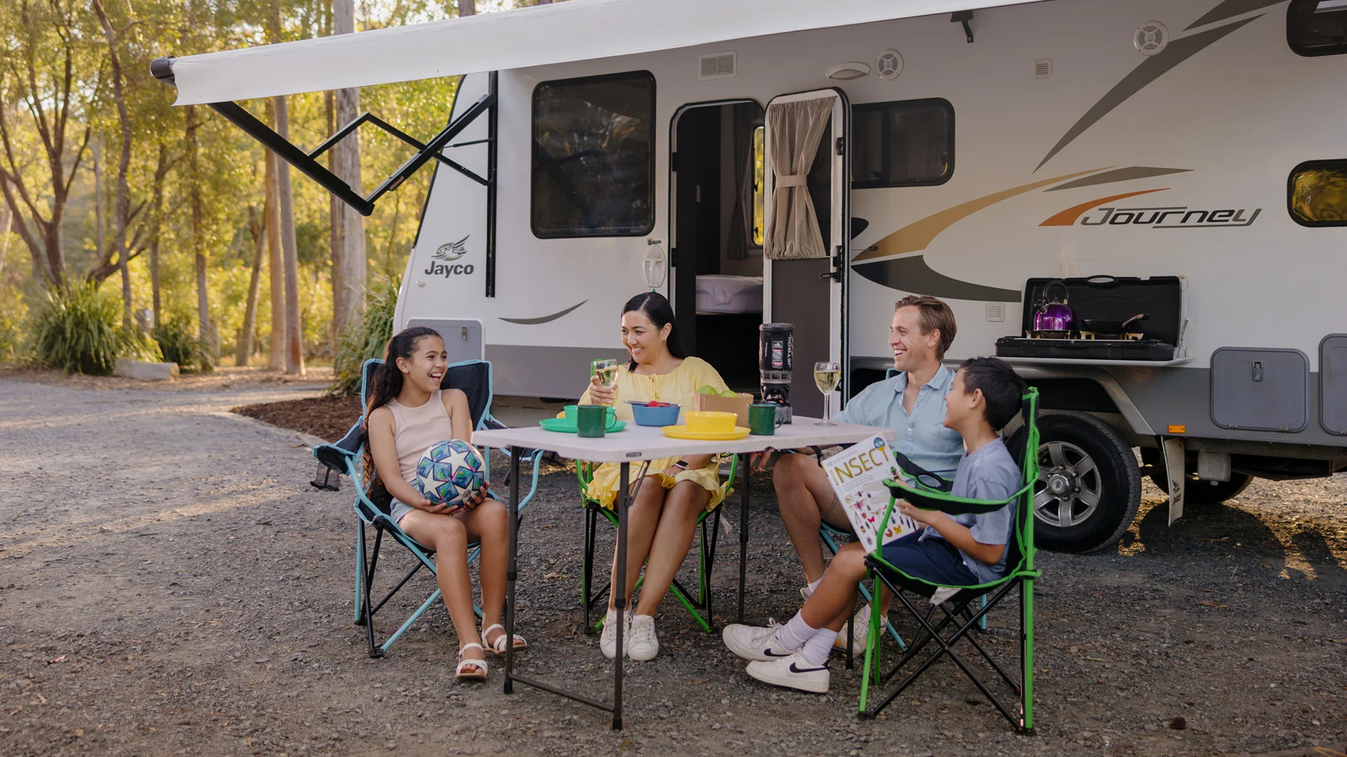 A family of four sits at a table outside their camper van, smiling and talking, with camping gear and trees in the background.