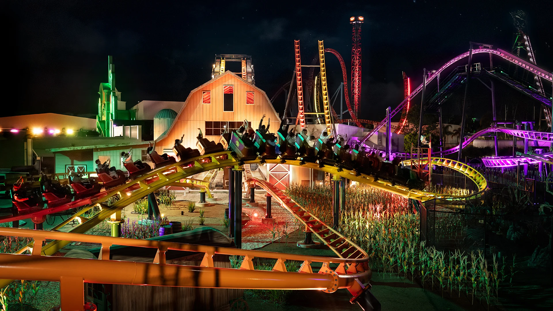 A brightly lit amusement park at night features a roller coaster full of riders, colorful lights illuminating the tracks and surrounding buildings, and other thrill rides visible in the background.